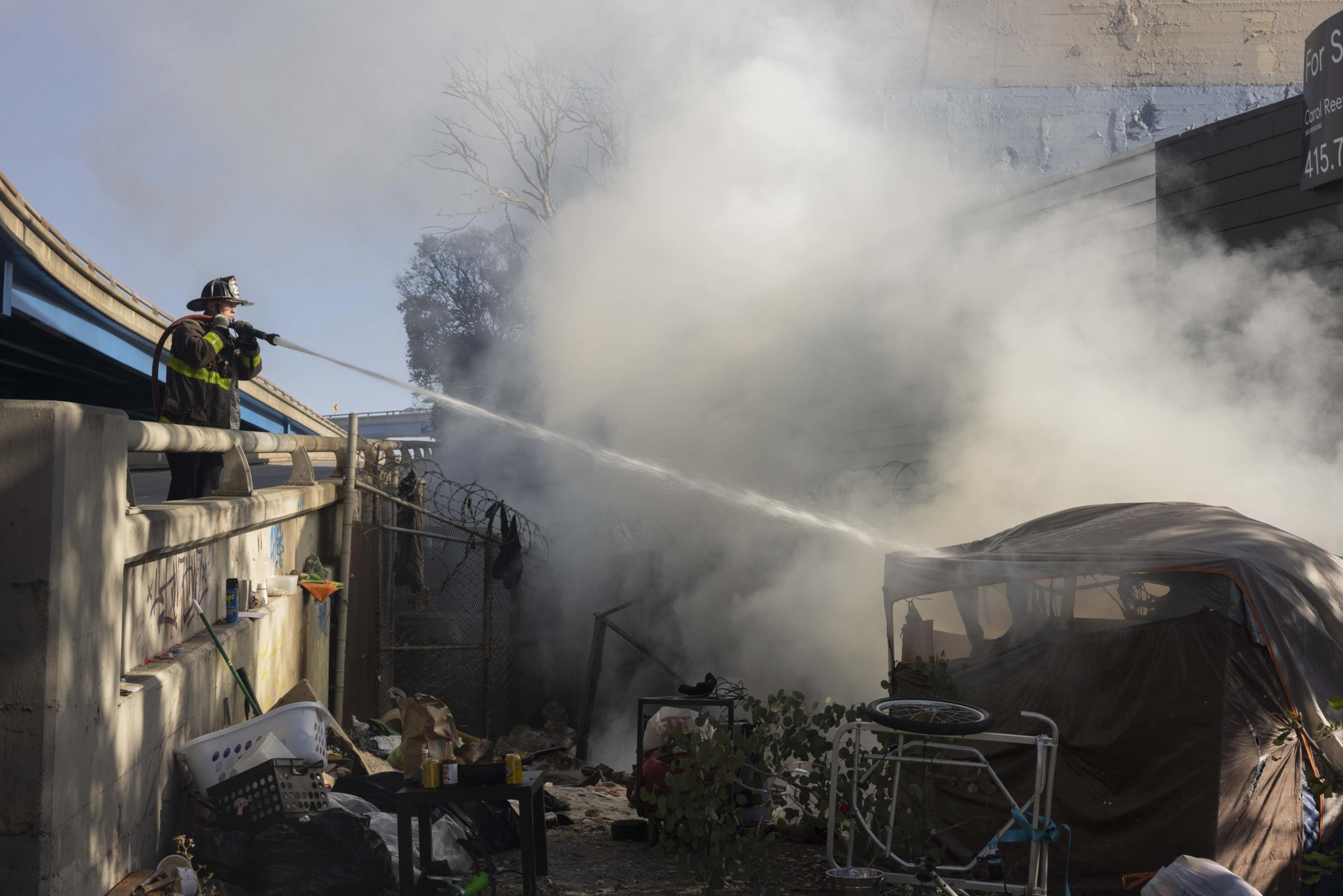 A firefighter sprays water onto a smoky area near a makeshift tent encampment under an overpass, surrounded by scattered items and debris.
