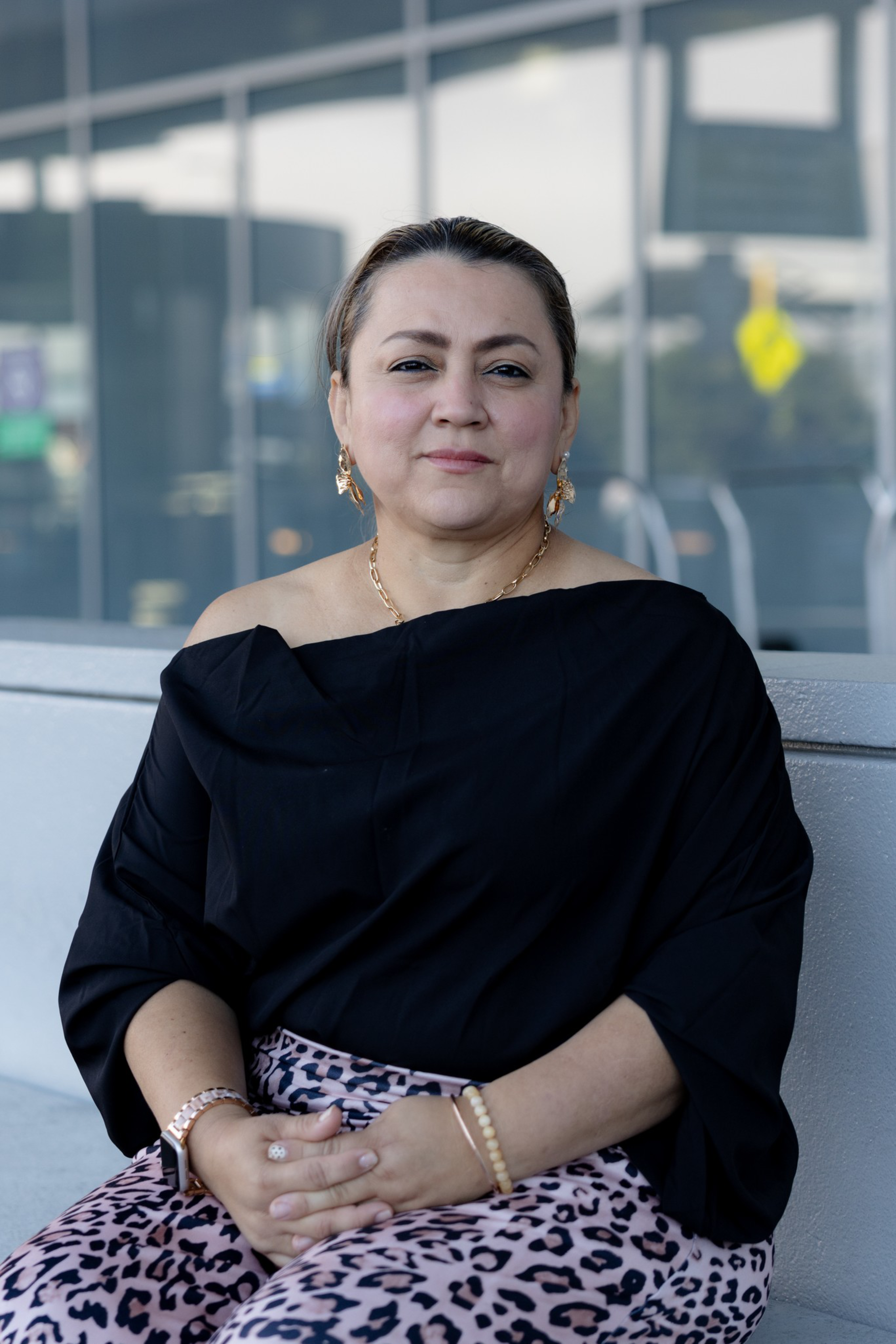 A woman with light makeup, wearing a black off-shoulder top and pink leopard-print pants, sits with hands clasped on her lap outdoors.