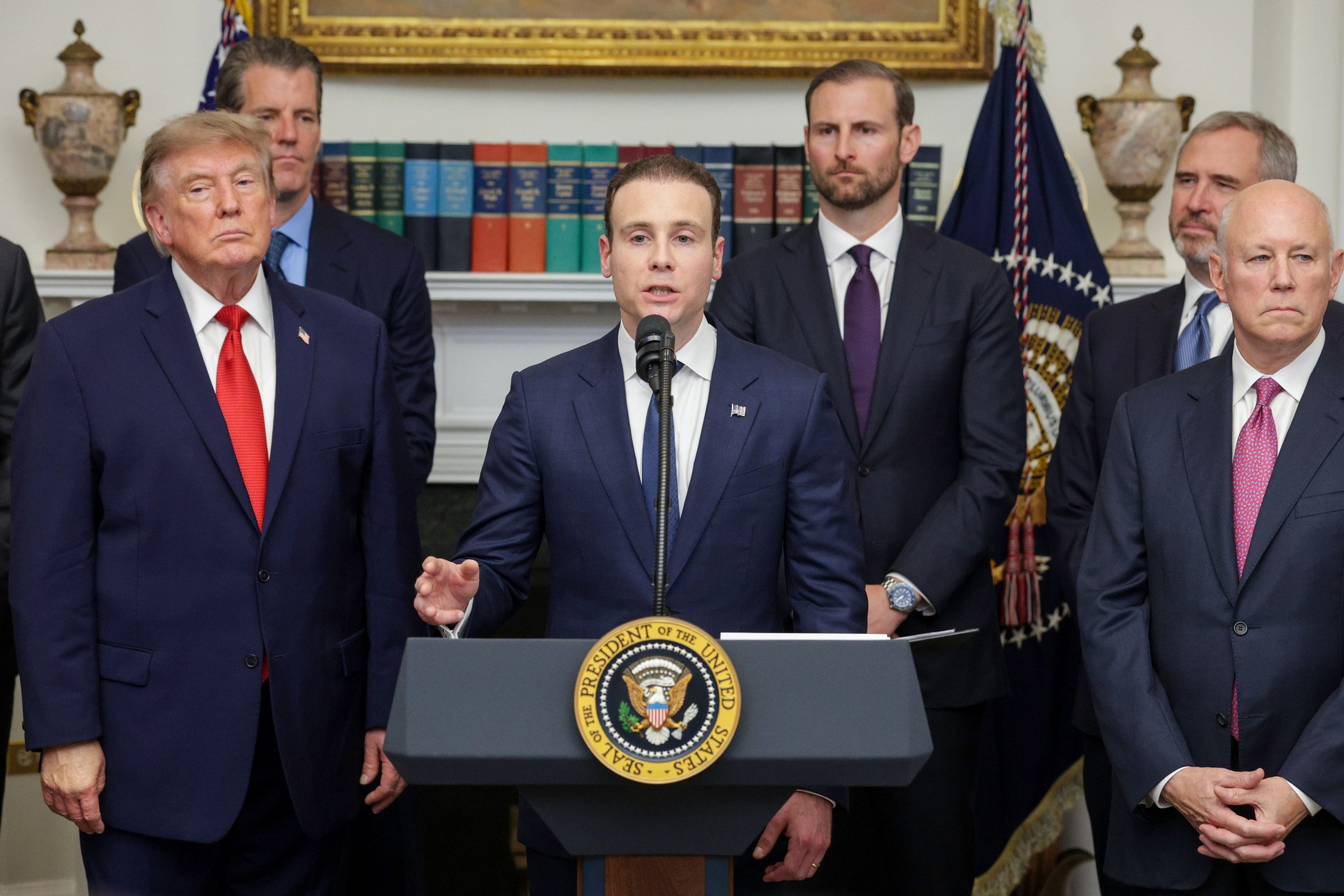 A man in a suit speaks at a podium with the presidential seal, flanked by five other men in suits standing in a formal room.