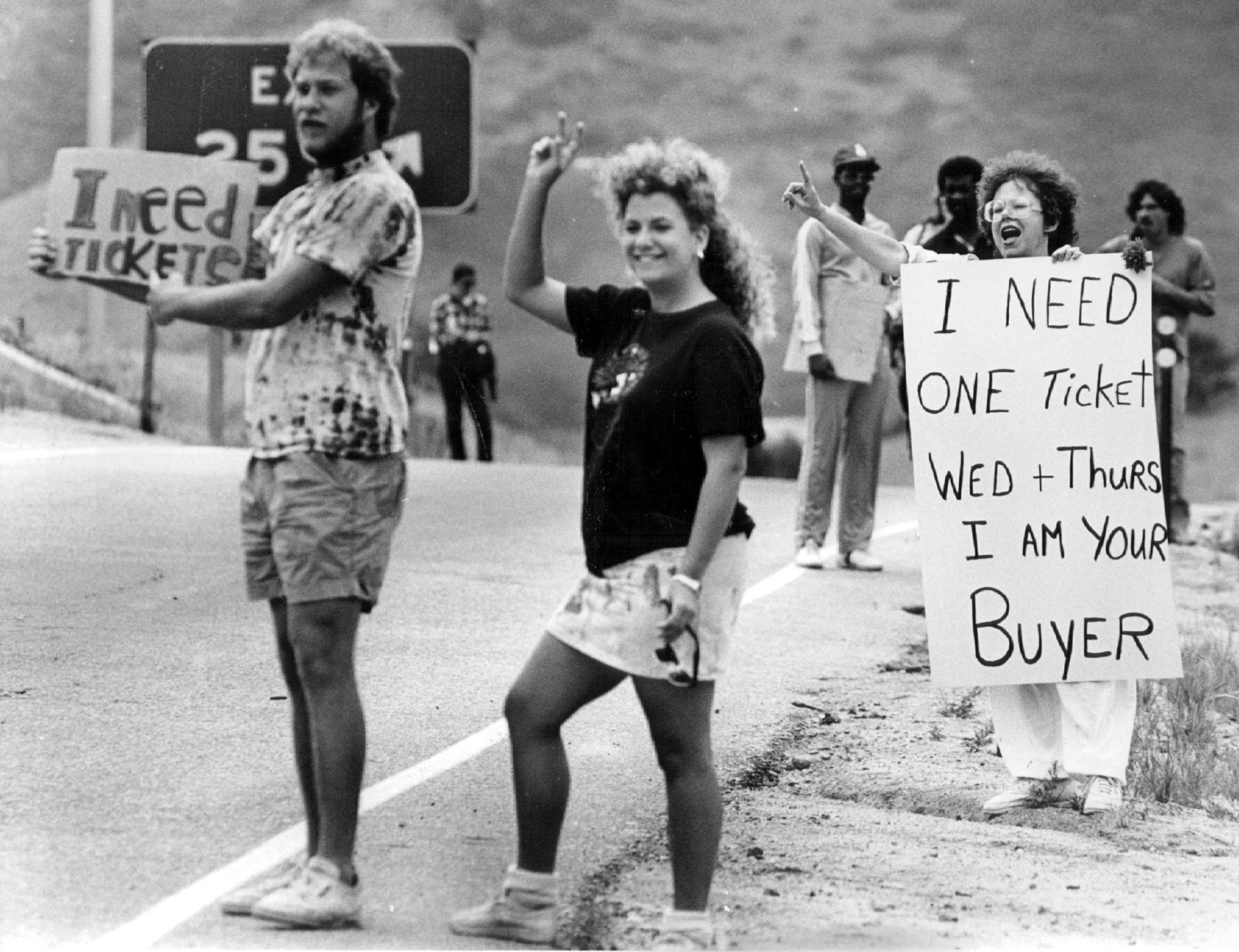 A group of people stand by a road holding signs seeking tickets, including one that reads &quot;I need tickets&quot; and another saying &quot;I need one ticket Wed + Thurs.&quot;