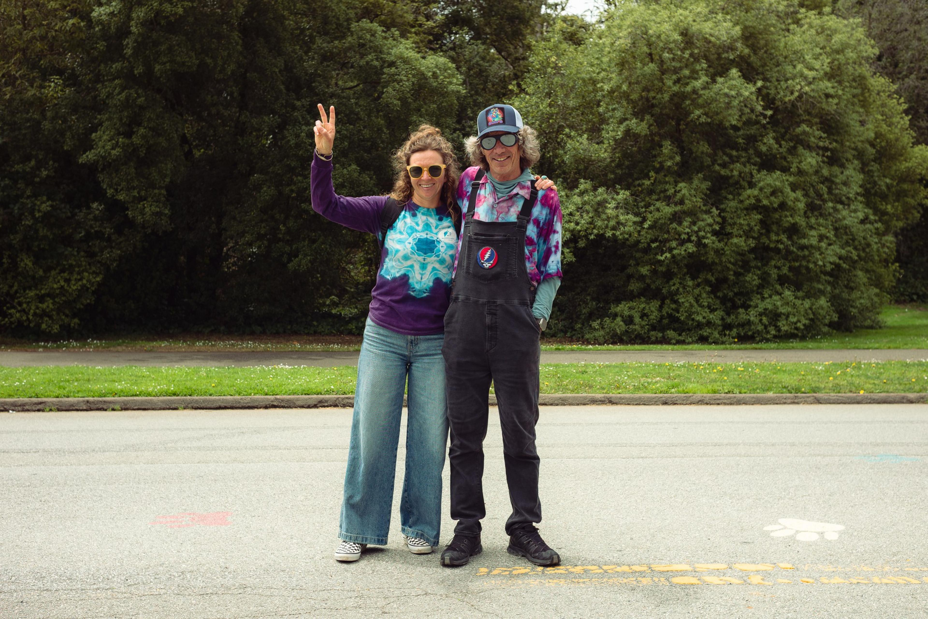 A man and woman stand on a street in casual tie-dye clothing, smiling, with the woman flashing a peace sign and trees in the background.