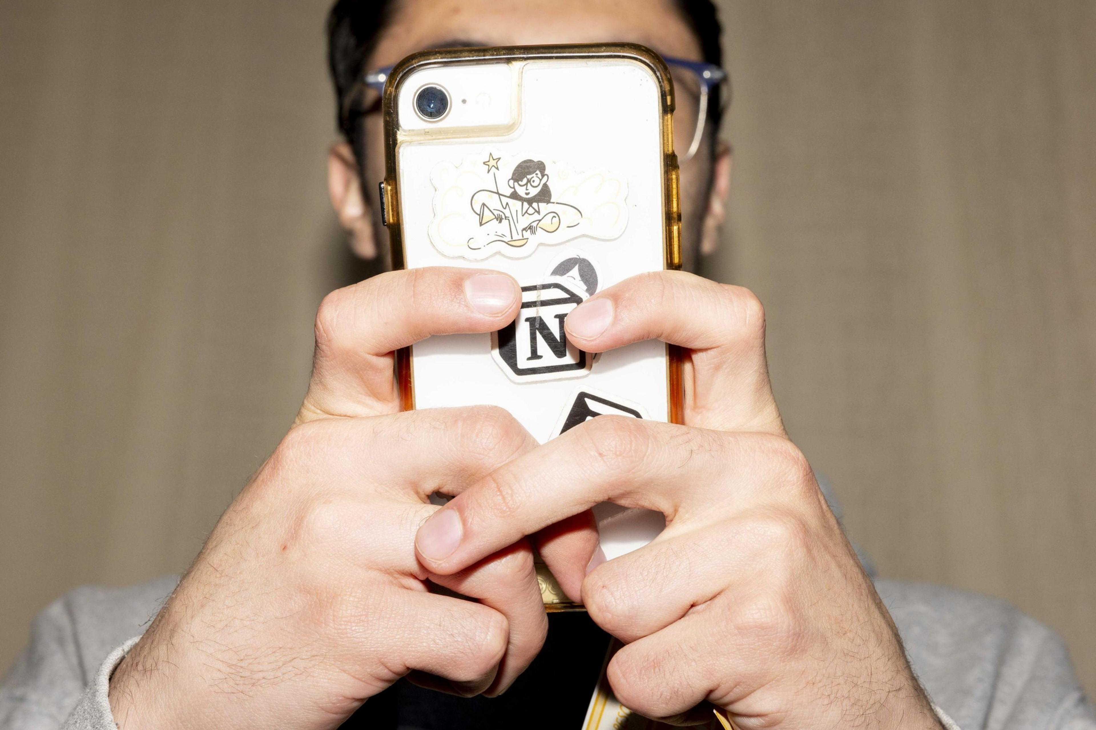 A person holds a phone with both hands, covering most of their face. The phone has a clear case displaying various playful stickers.