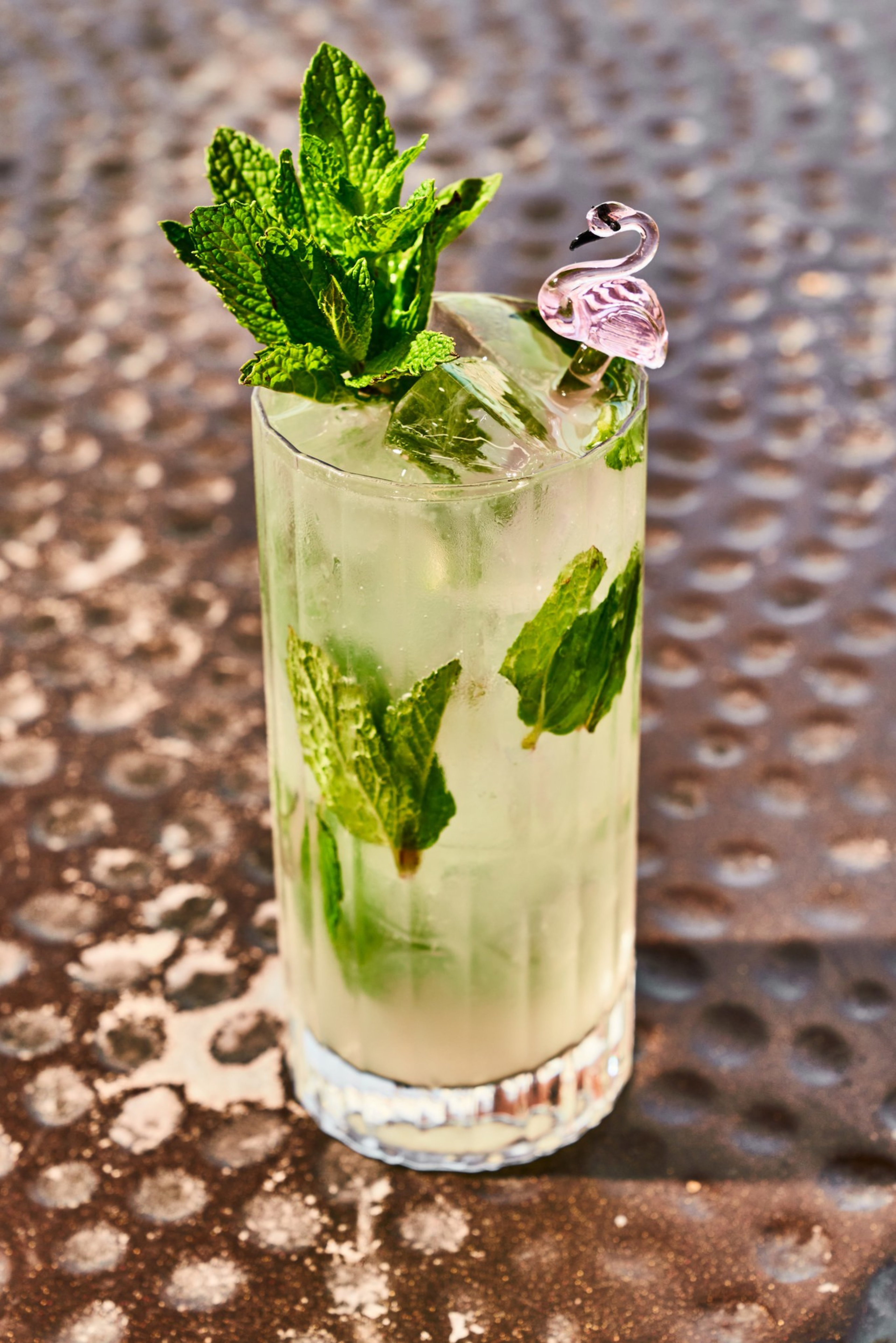 A tall glass filled with a refreshing minty drink, garnished with fresh mint leaves and a small pink flamingo cocktail stick, sits on a textured table.