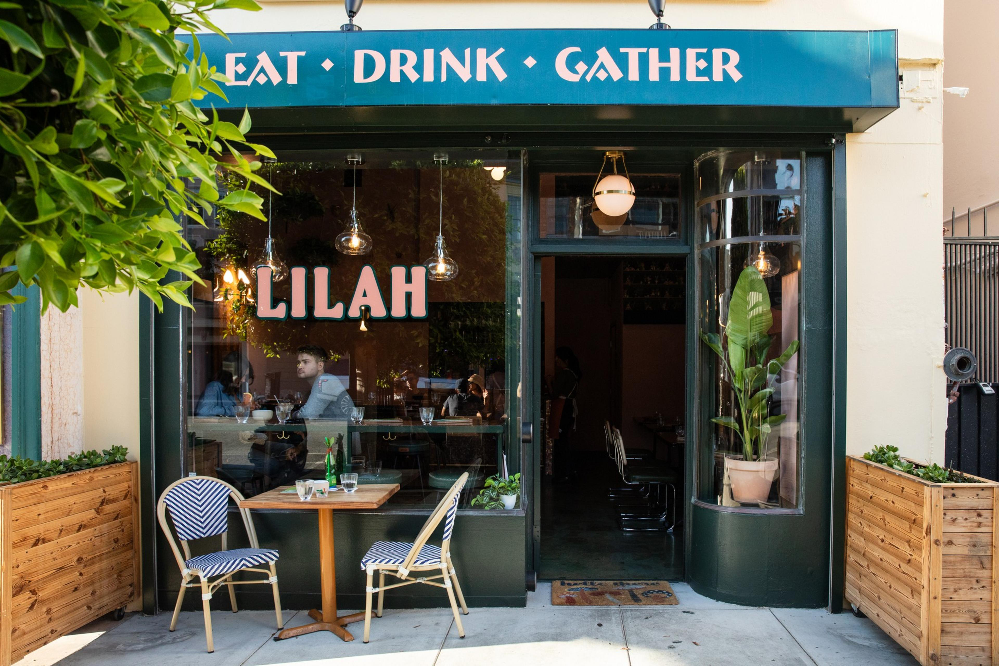A charming café entrance with a green facade, displaying &quot;Eat, Drink, Gather.&quot; Two small tables with chairs sit outside, flanked by potted plants. Inside, patrons are dining.