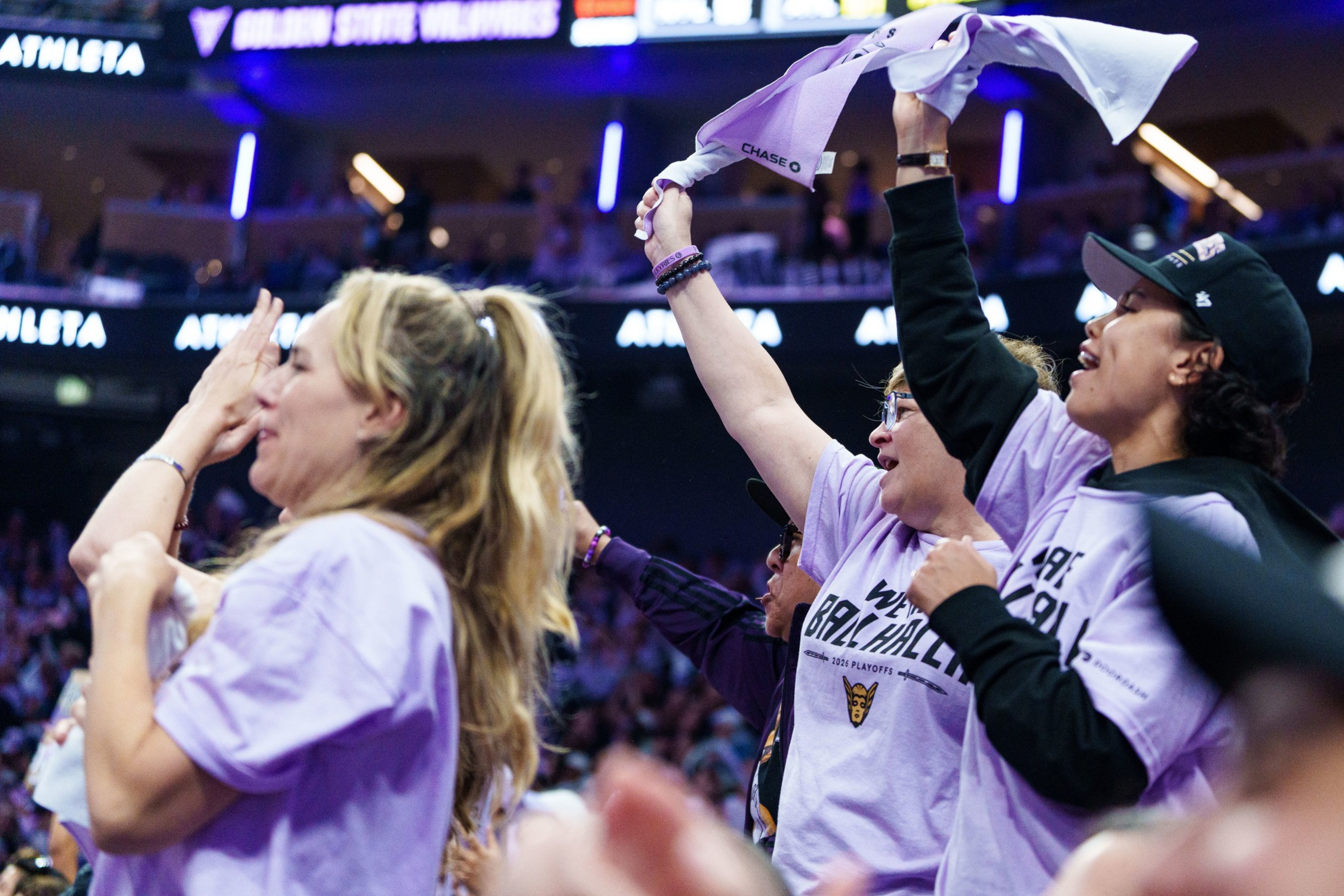 Three enthusiastic fans wearing matching lavender shirts cheer and wave towels during a lively sports event.