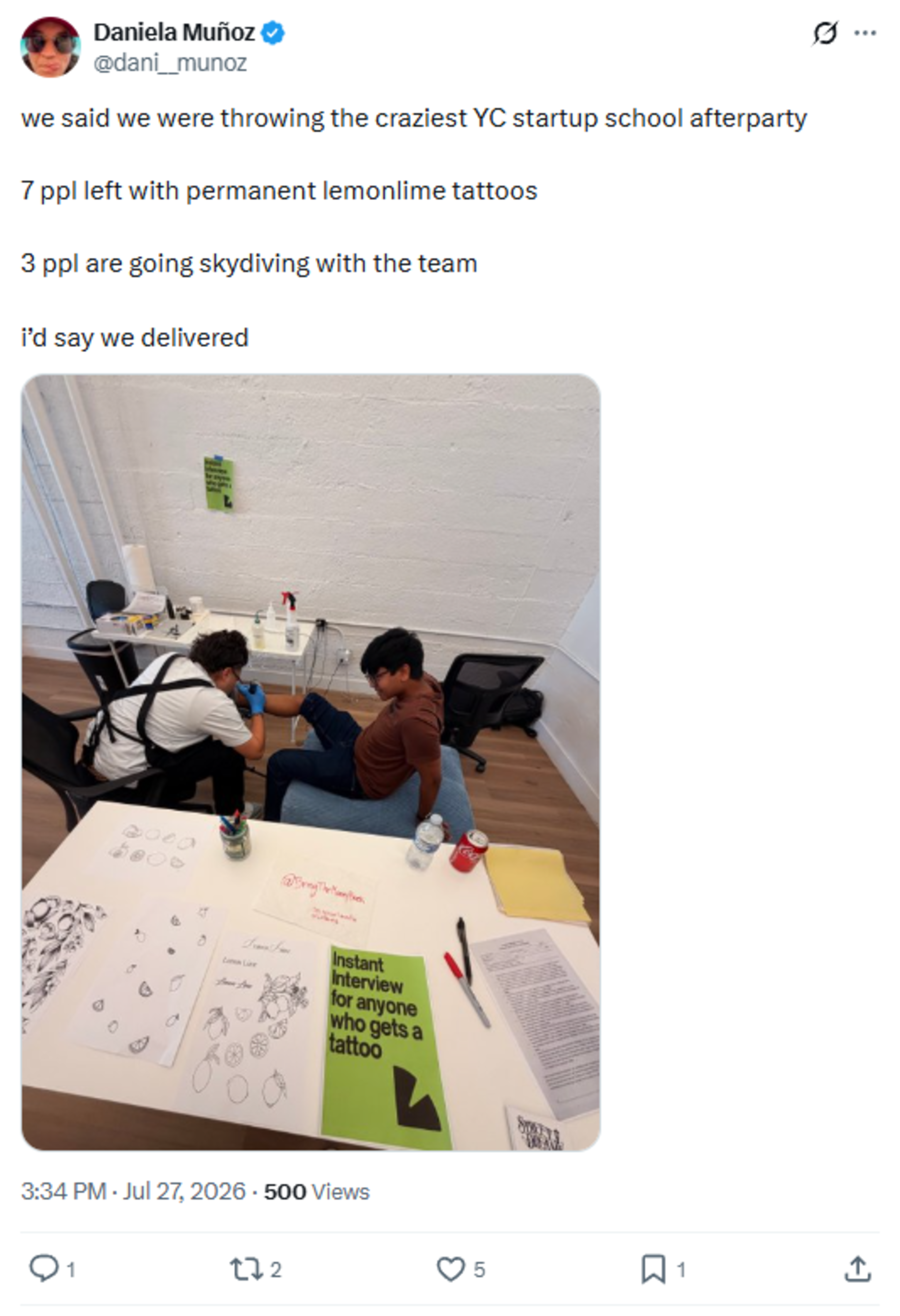 A person is getting a tattoo on their leg by an artist in a white room, with tattoo design sheets and papers spread on a table nearby.