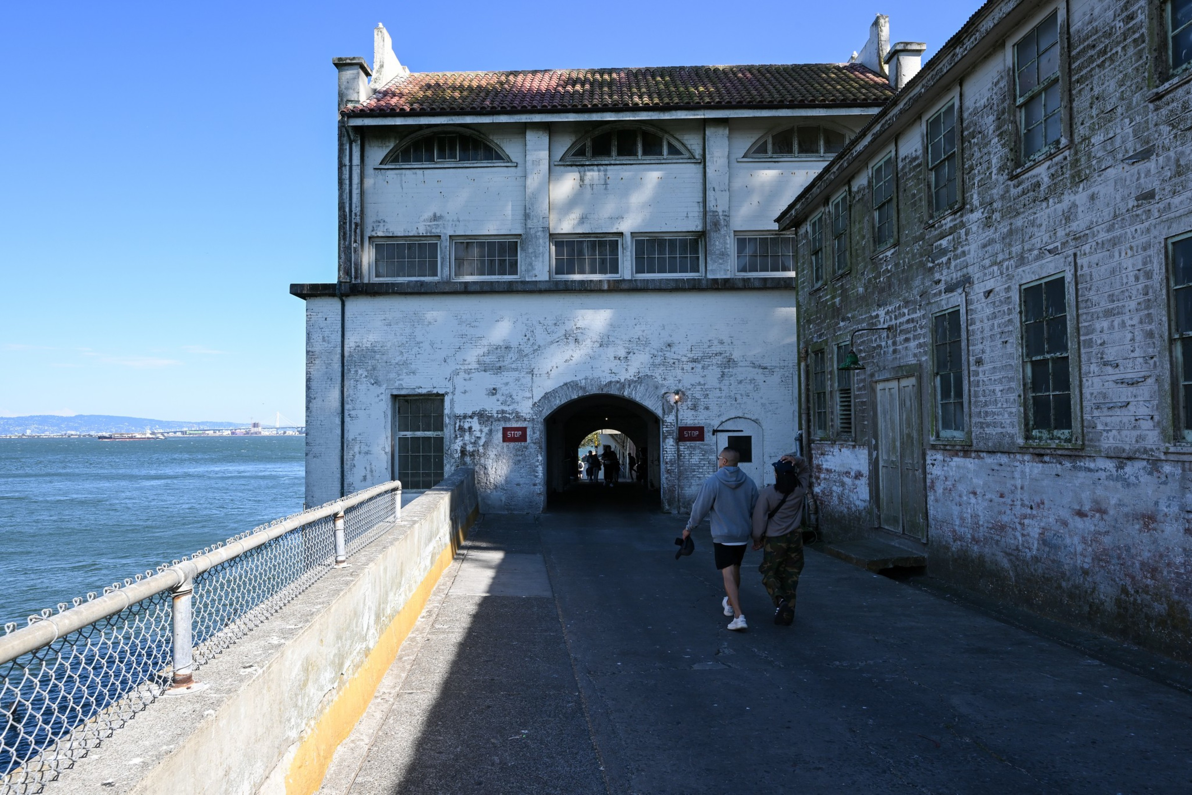 A couple walks towards an arched entryway between two weathered buildings by the water, with a distant view of a bridge under a clear blue sky.