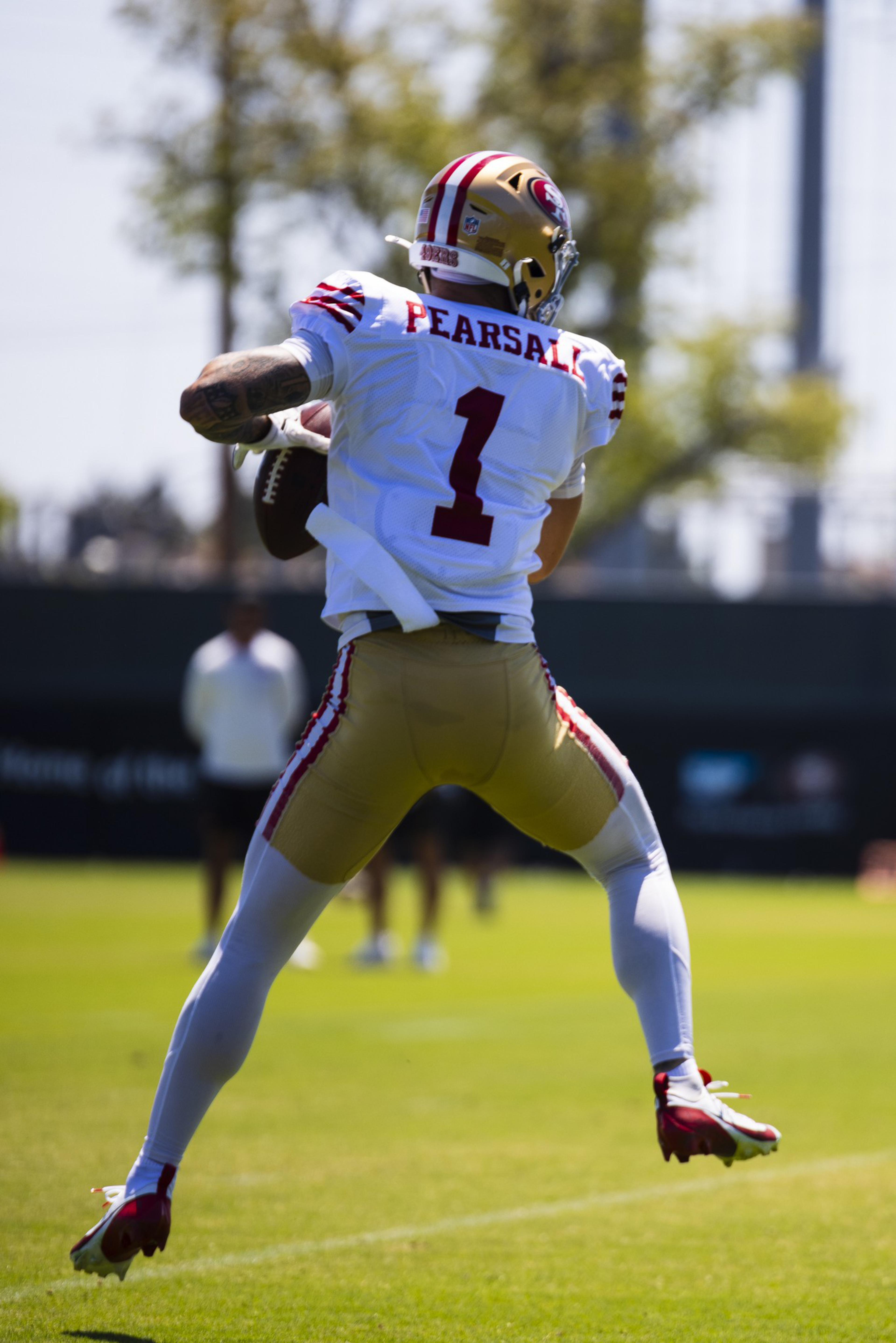 A football player wearing a white jersey with "PEARSALL" and number 1 is airborne, holding the ball as he prepares to throw it.