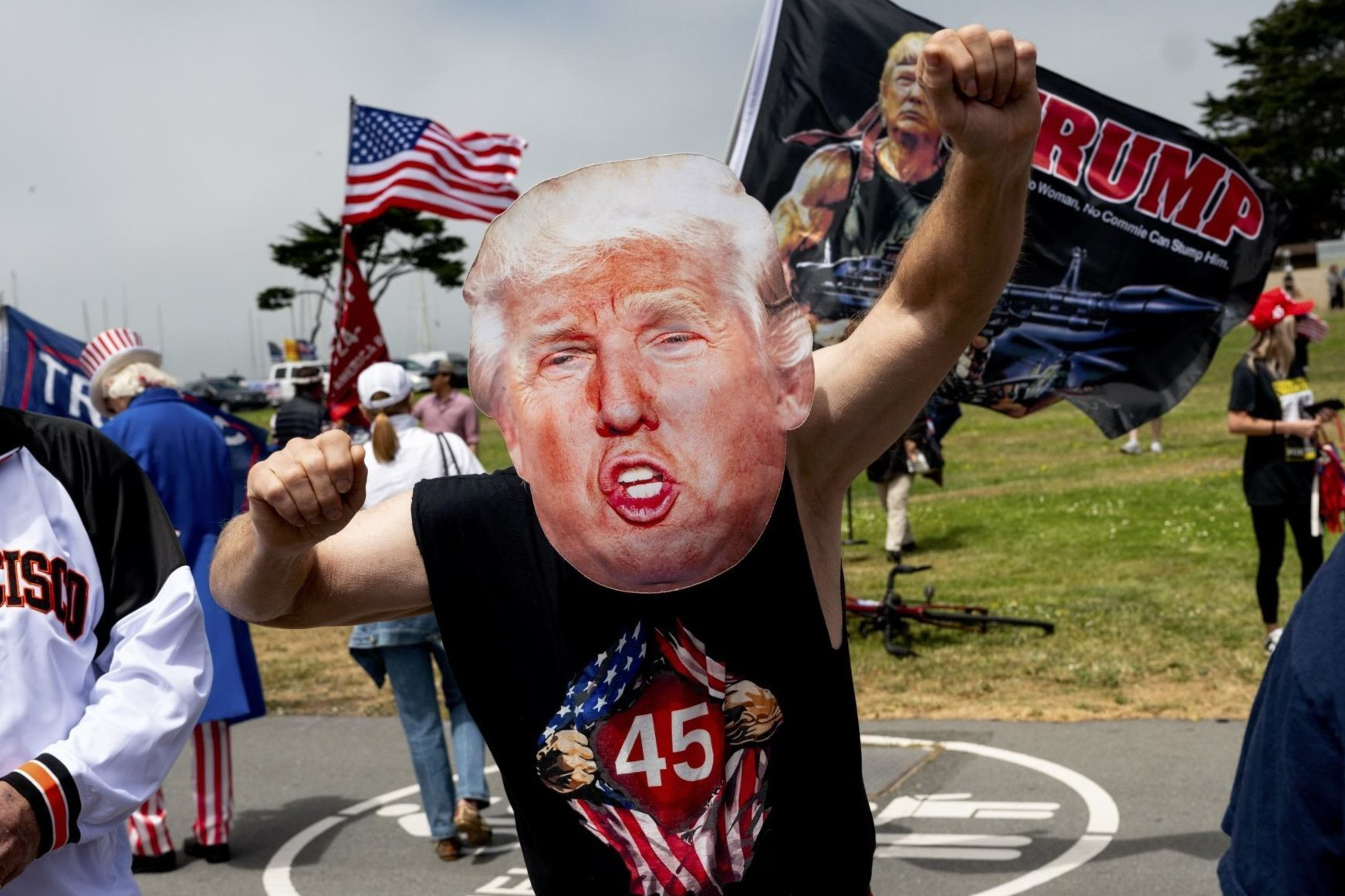 A person wearing a Donald Trump mask is flexing their arms in front of a group of people holding American flags and "Trump" banners. Other attendees are also visible.