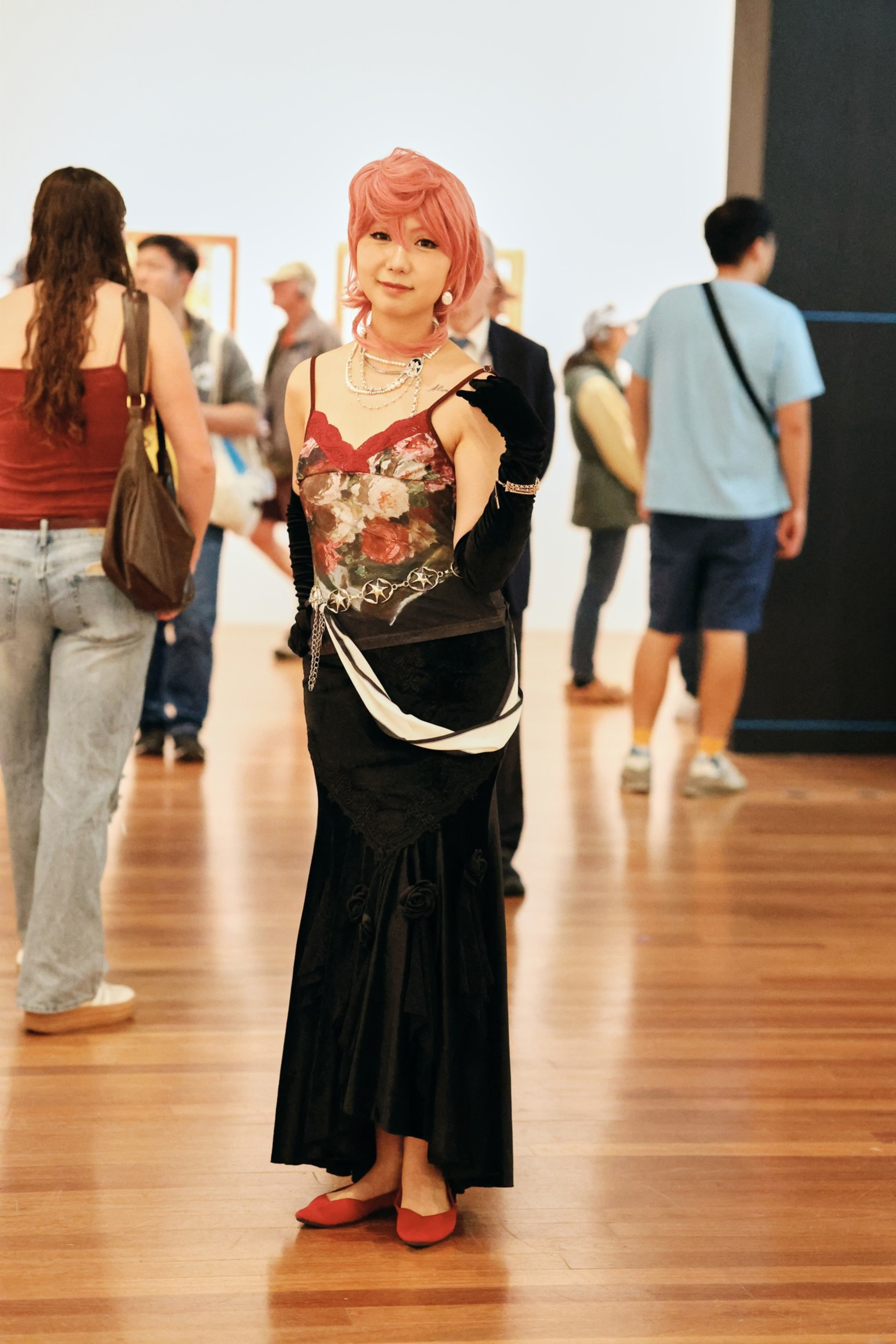 A person with pink hair wears a floral top, black long skirt, gloves, red shoes, and silver jewelry, standing on a wooden floor with people around.