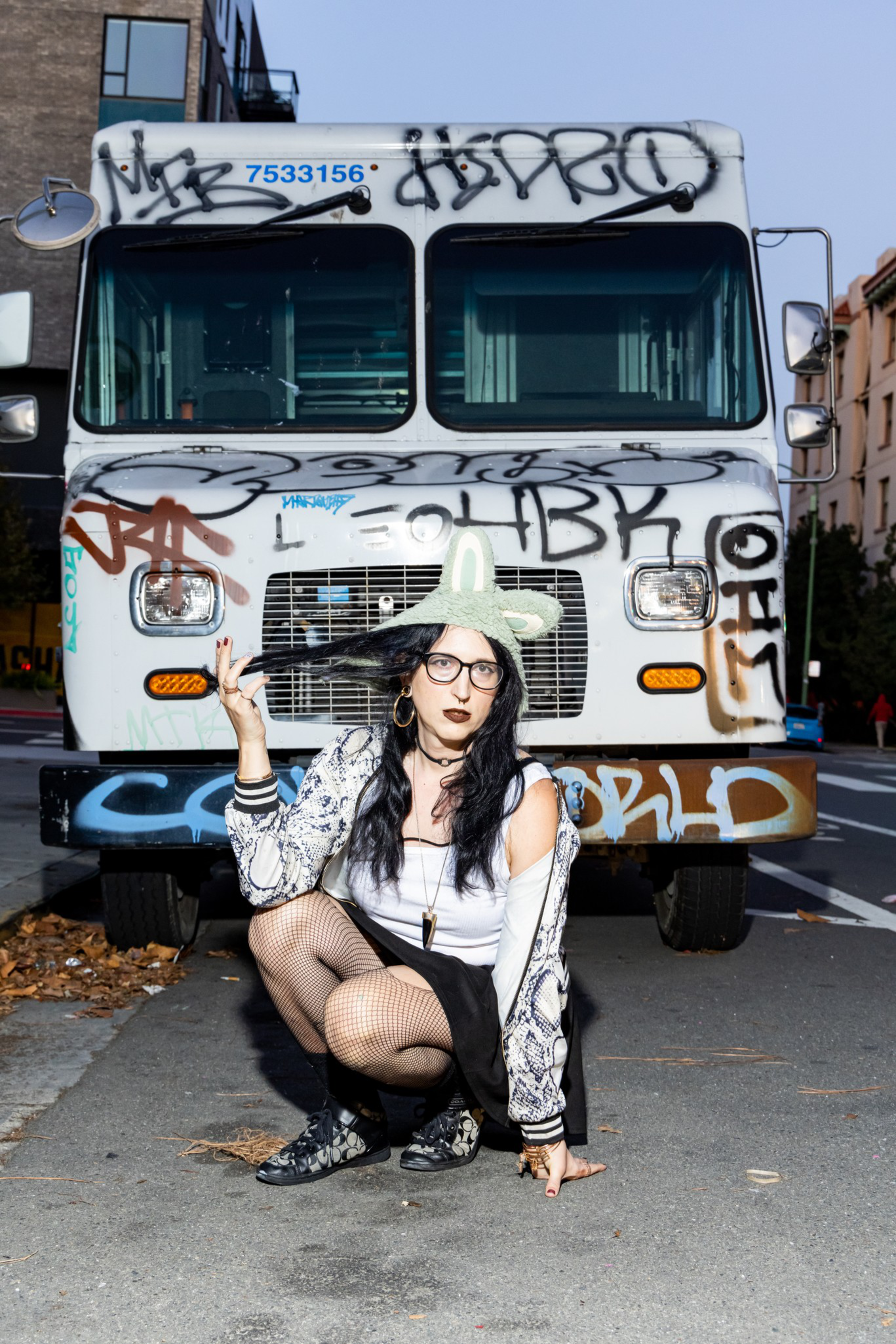A woman with long black hair, glasses, and a green hat squats in front of a graffiti-covered white truck, holding a strand of her hair with one hand.