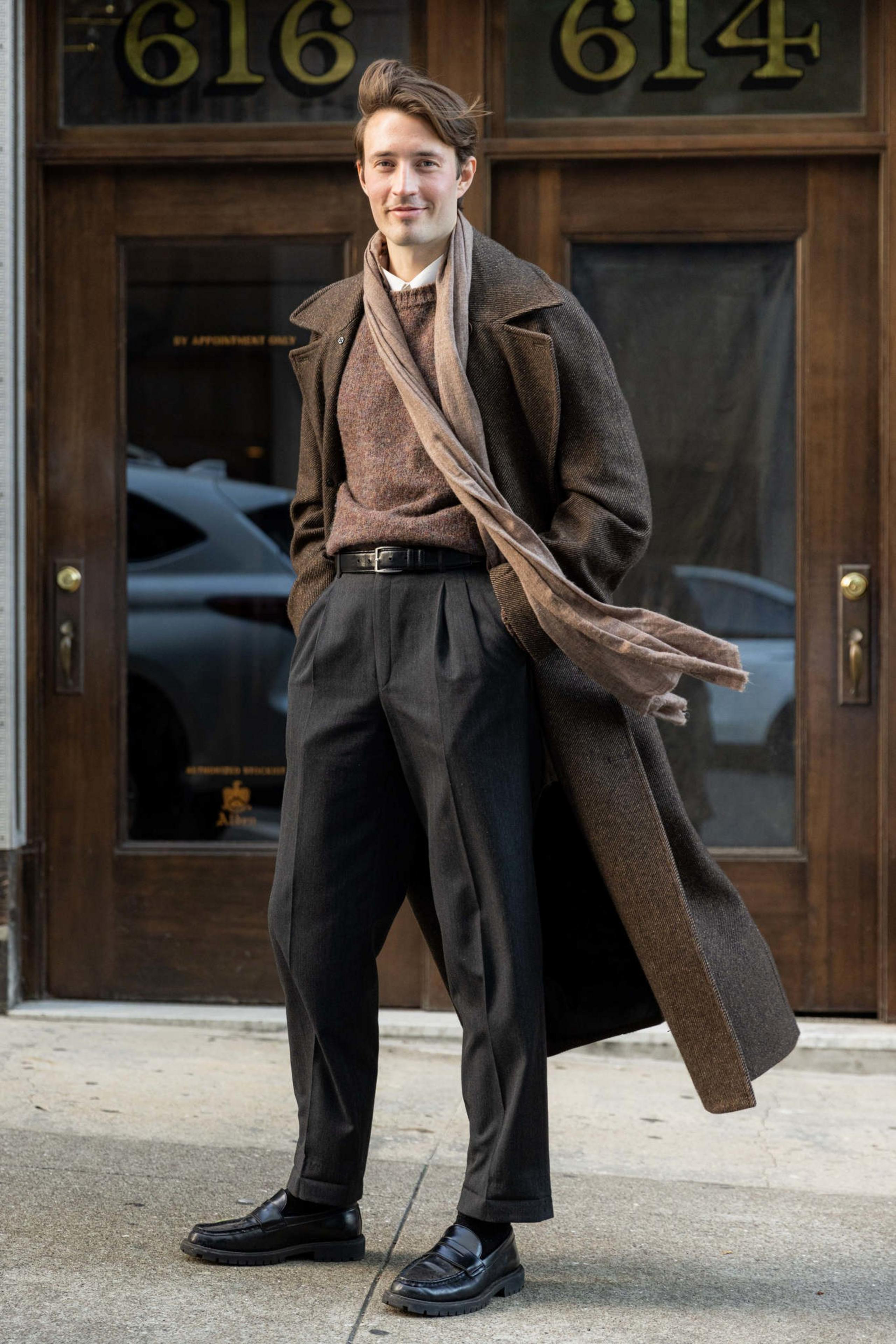 A man stands outside a building wearing a brown overcoat, brown sweater, light scarf, black trousers, and black shoes, smiling with hands in pockets.