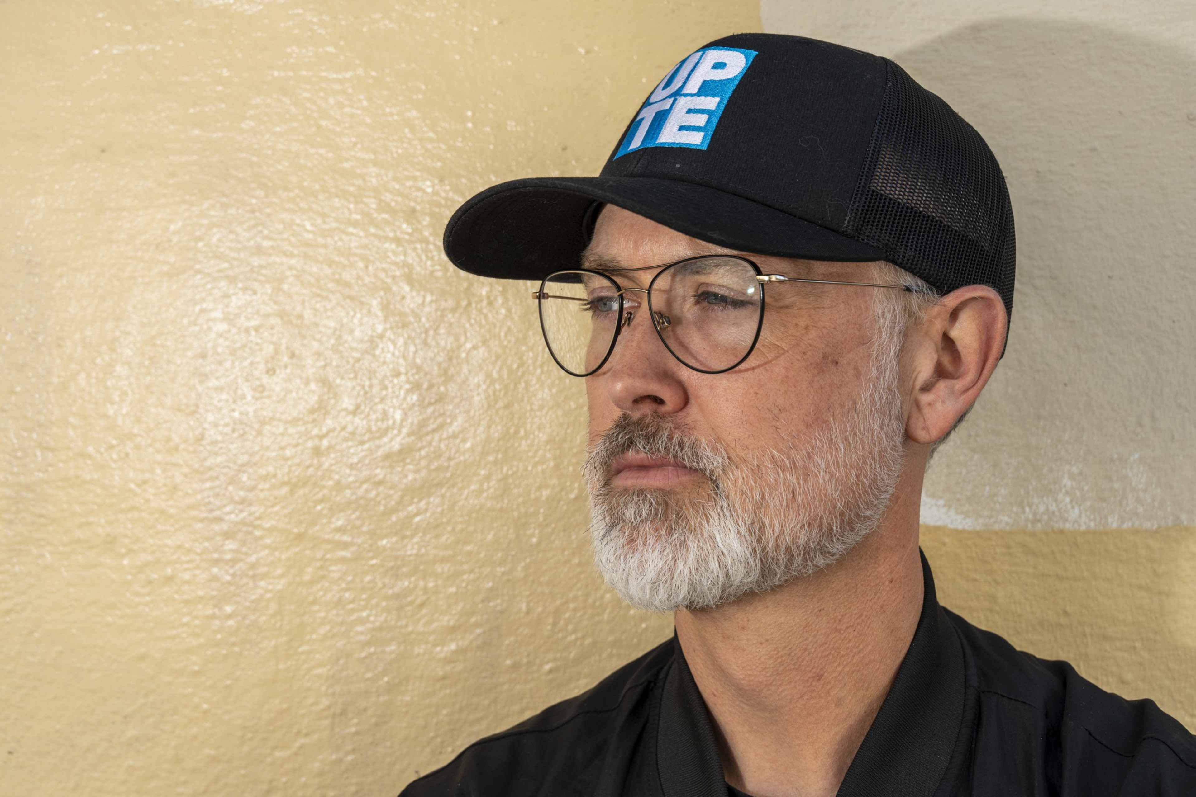 A man with a gray beard and glasses wears a black cap with blue and white text, looking to the side against a beige wall.