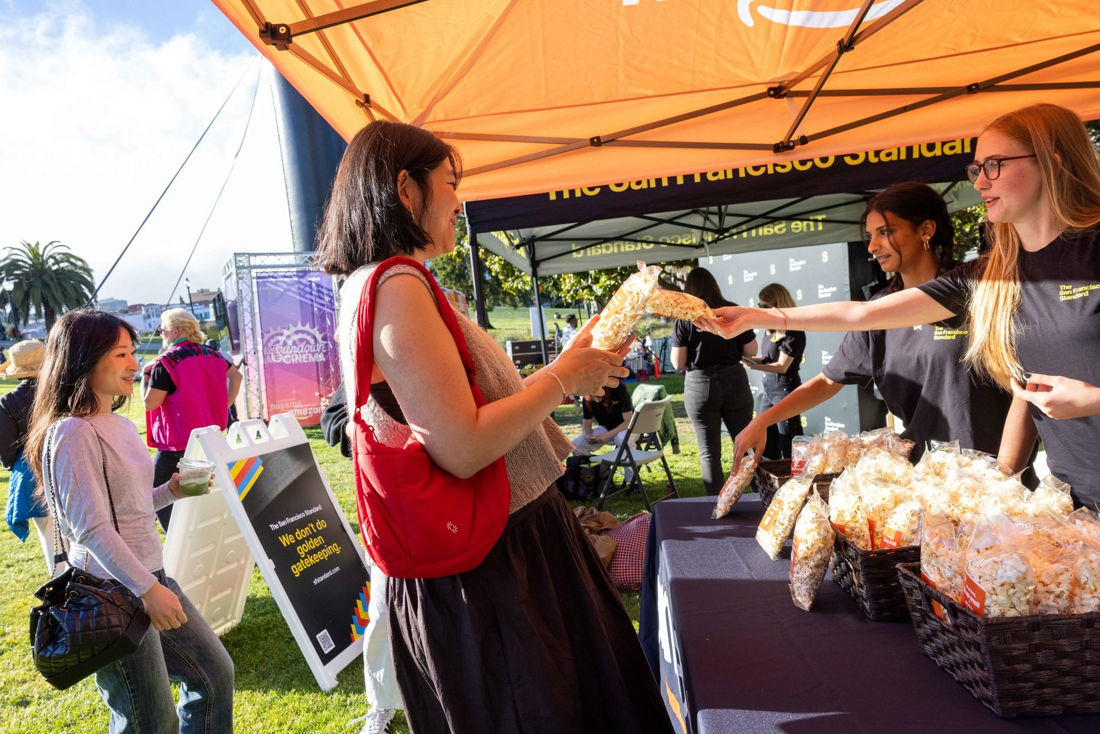 People are buying bags of popcorn at an outdoor event booth under an orange canopy, with several baskets of popcorn on the table.