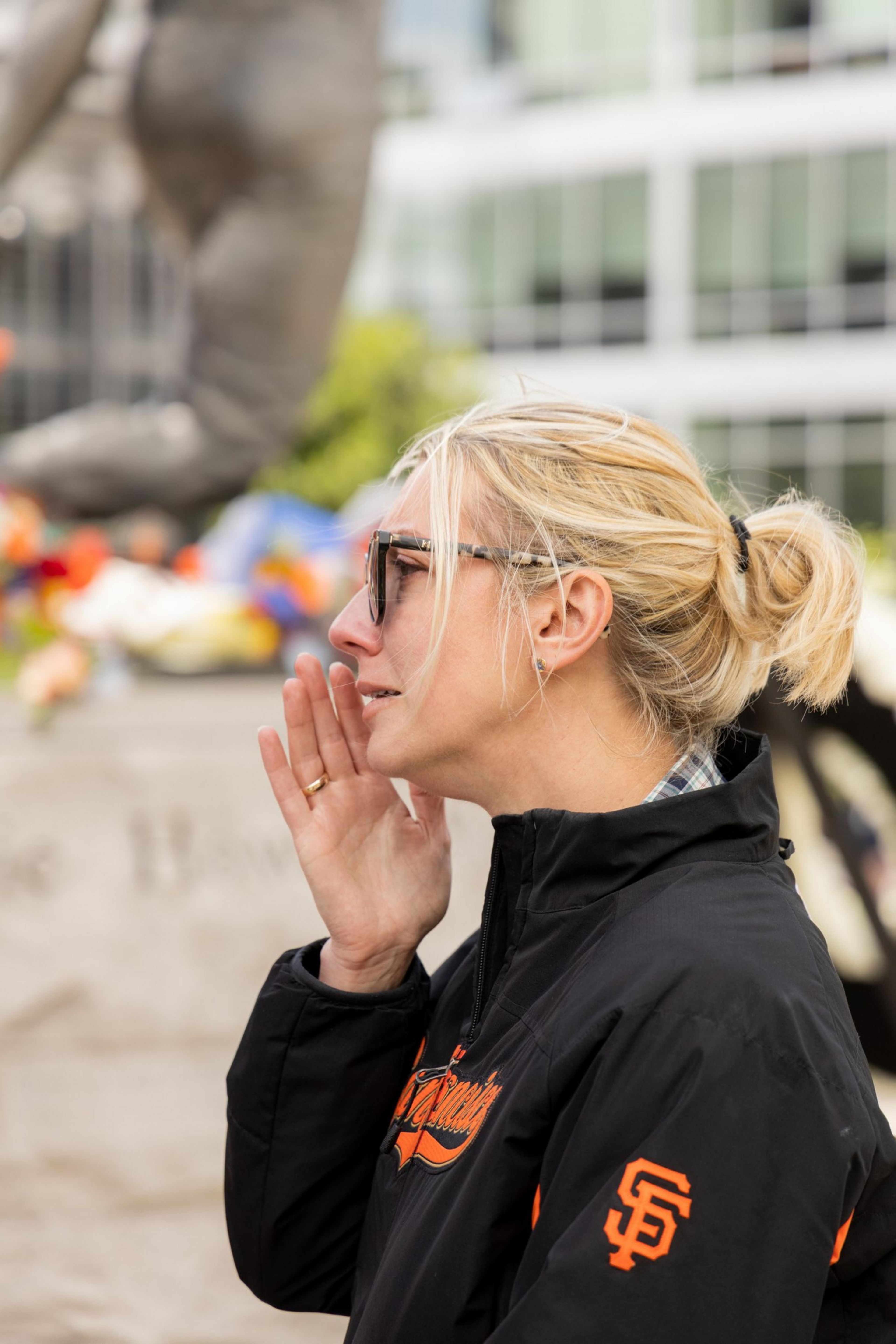 A blonde woman wearing sunglasses and a black San Francisco Giants jacket appears to be crying, with her hand up to her mouth, set against a blurred cityscape background.