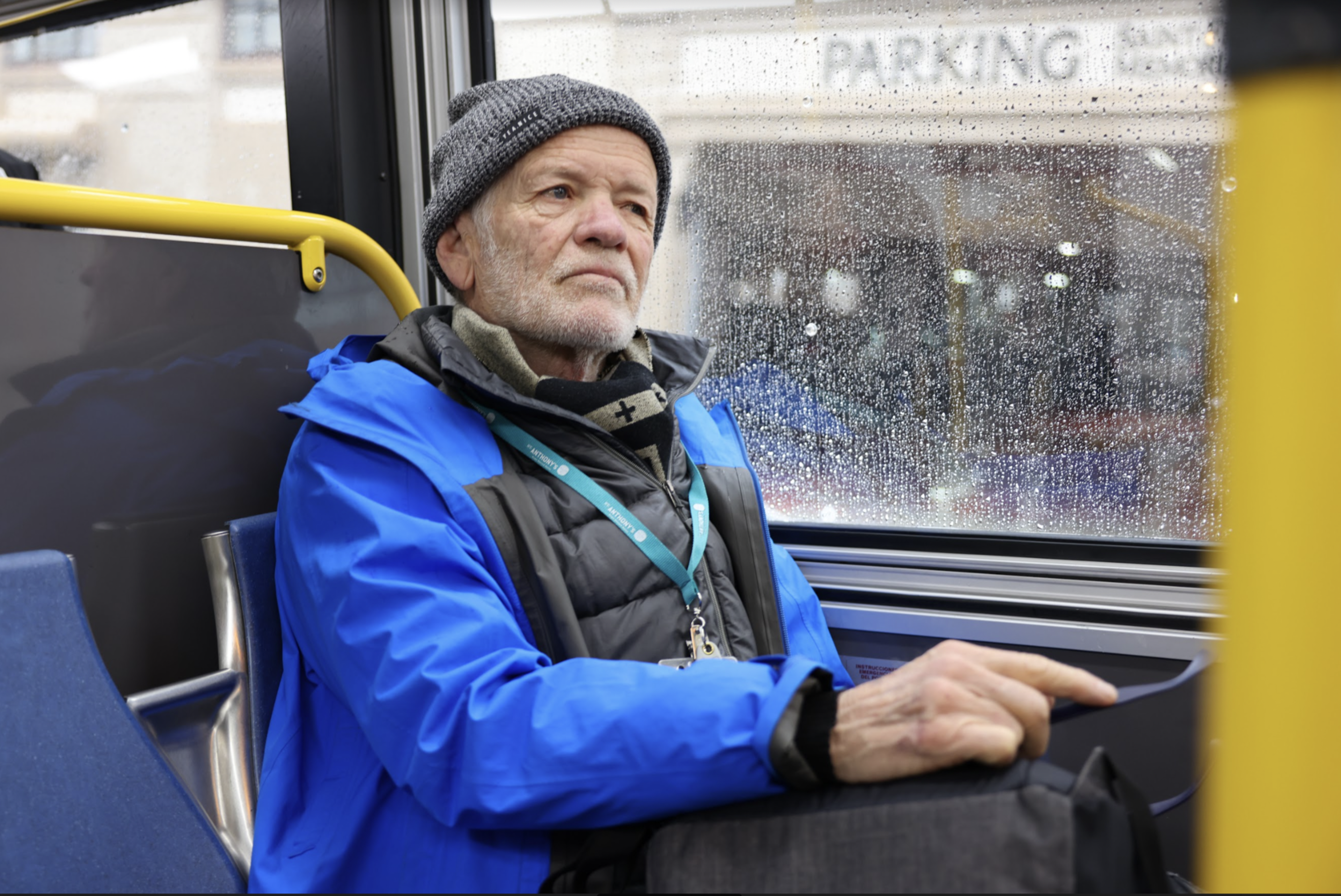 An elderly man wearing a blue jacket and gray beanie looks out a rain-speckled bus window, resting his hand on a black bag in his lap.