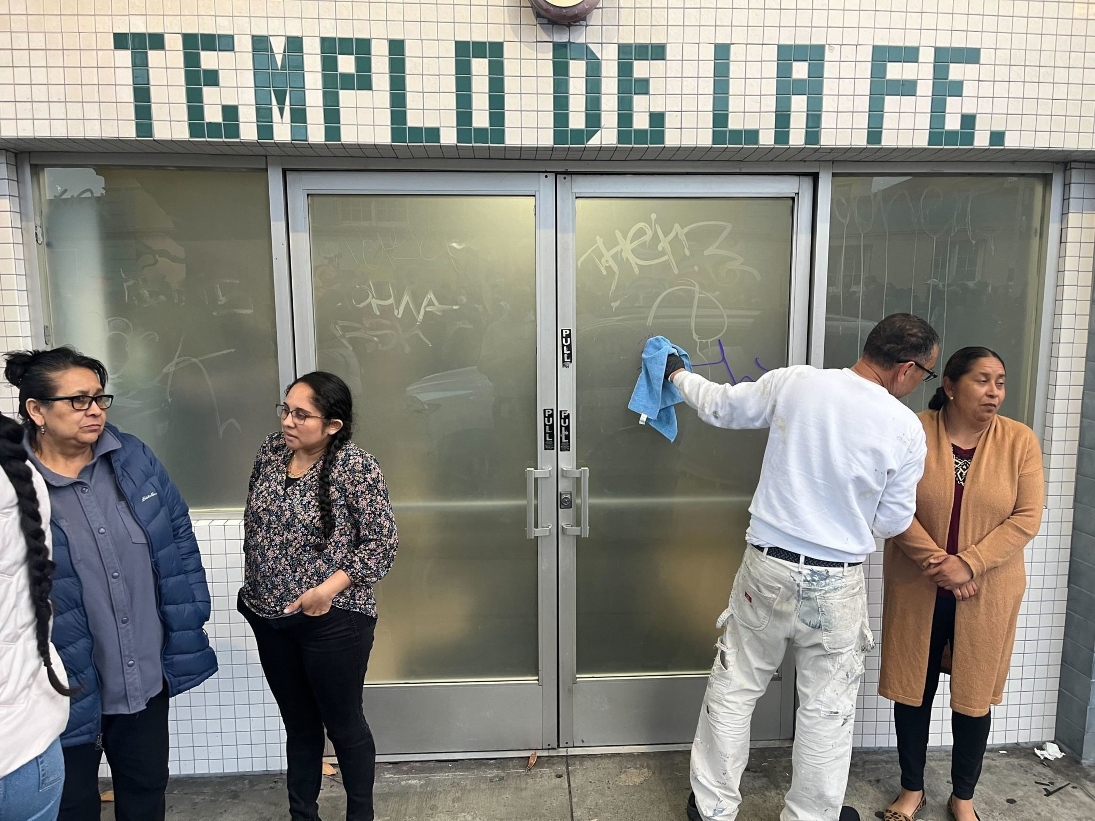 Four people stand outside a building labeled &quot;Templo de la Fe.&quot; One person is cleaning the glass door covered in graffiti, while the others stand nearby.