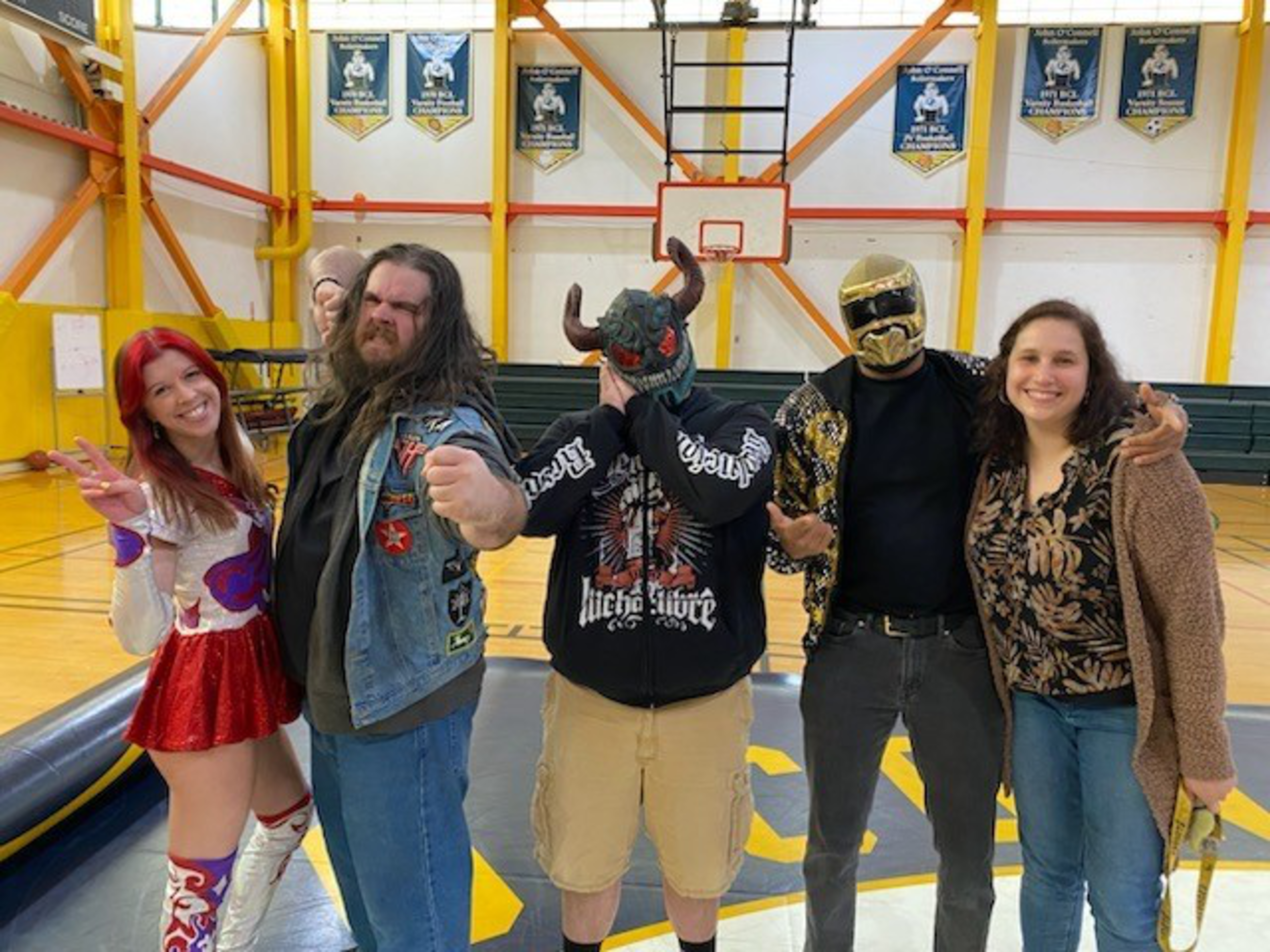 Five people stand in a gym, two wearing masks and one in a cheerleader outfit, posing with fists raised or arms around each other, smiling and playful.