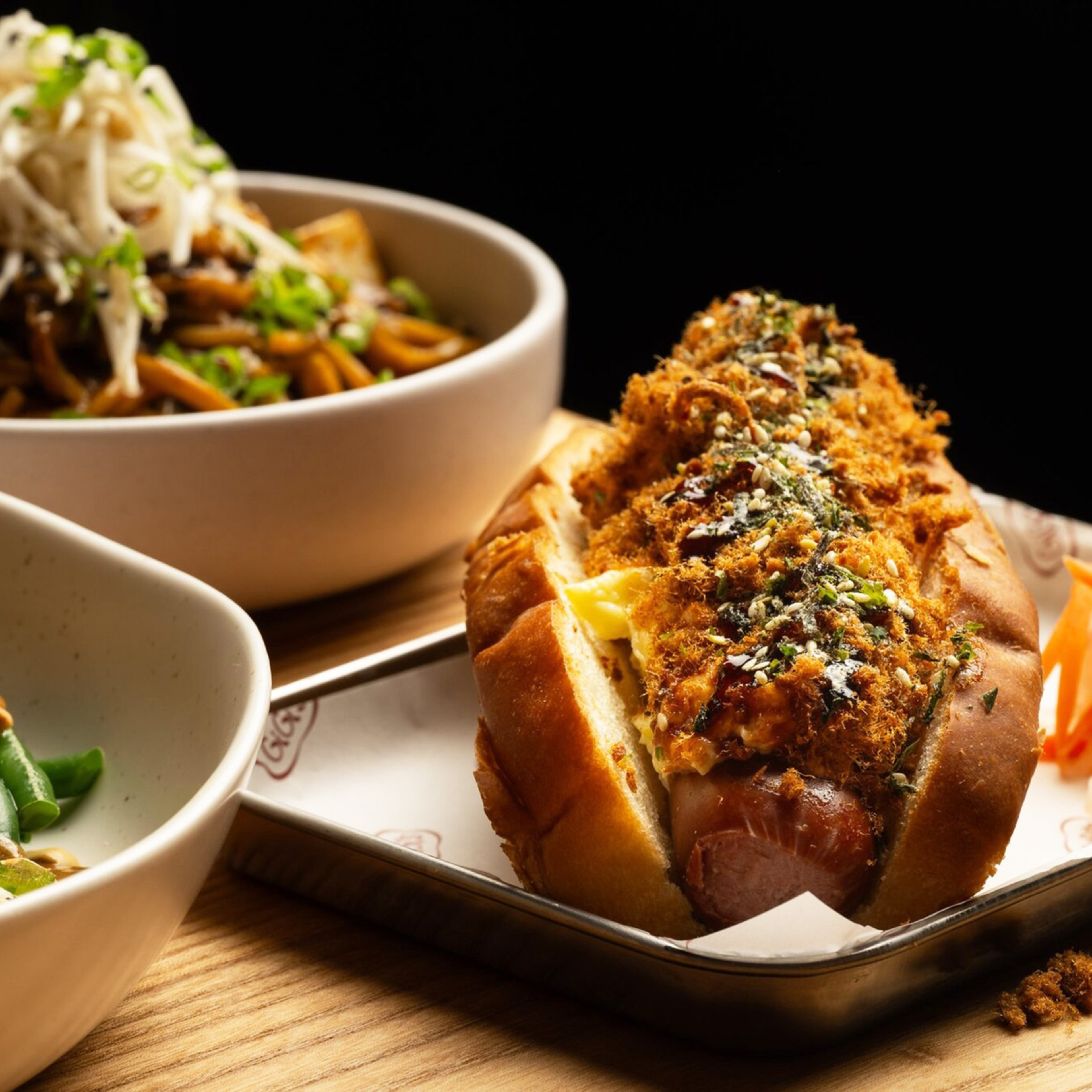 The image shows a plated hot dog topped with herbs and crispy bits, alongside bowls of noodles with sprouts and salad with radish and green beans.
