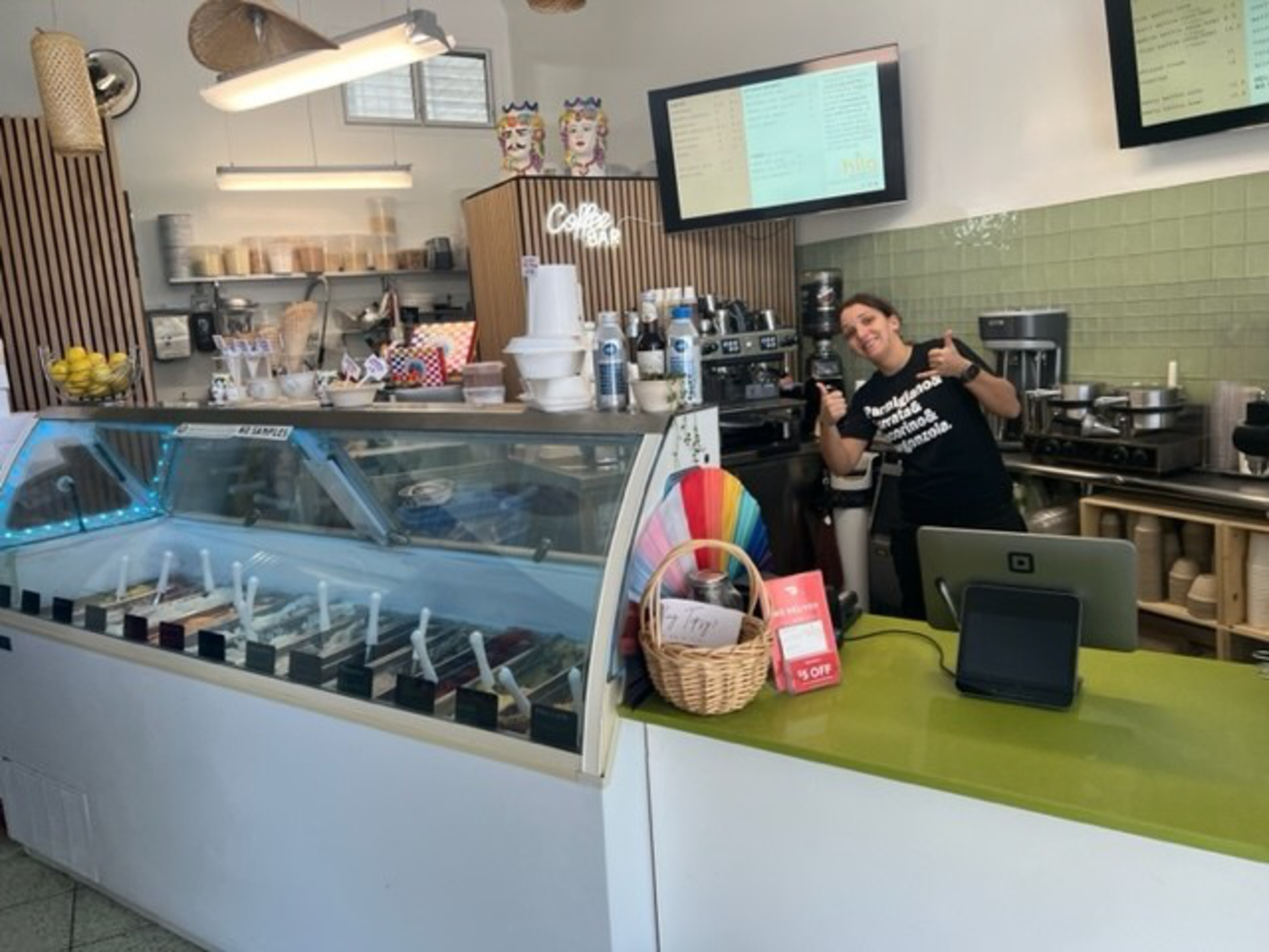 A smiling barista gives thumbs up behind a counter with a gelato display, coffee machines, and menu screens in a bright, modern café.