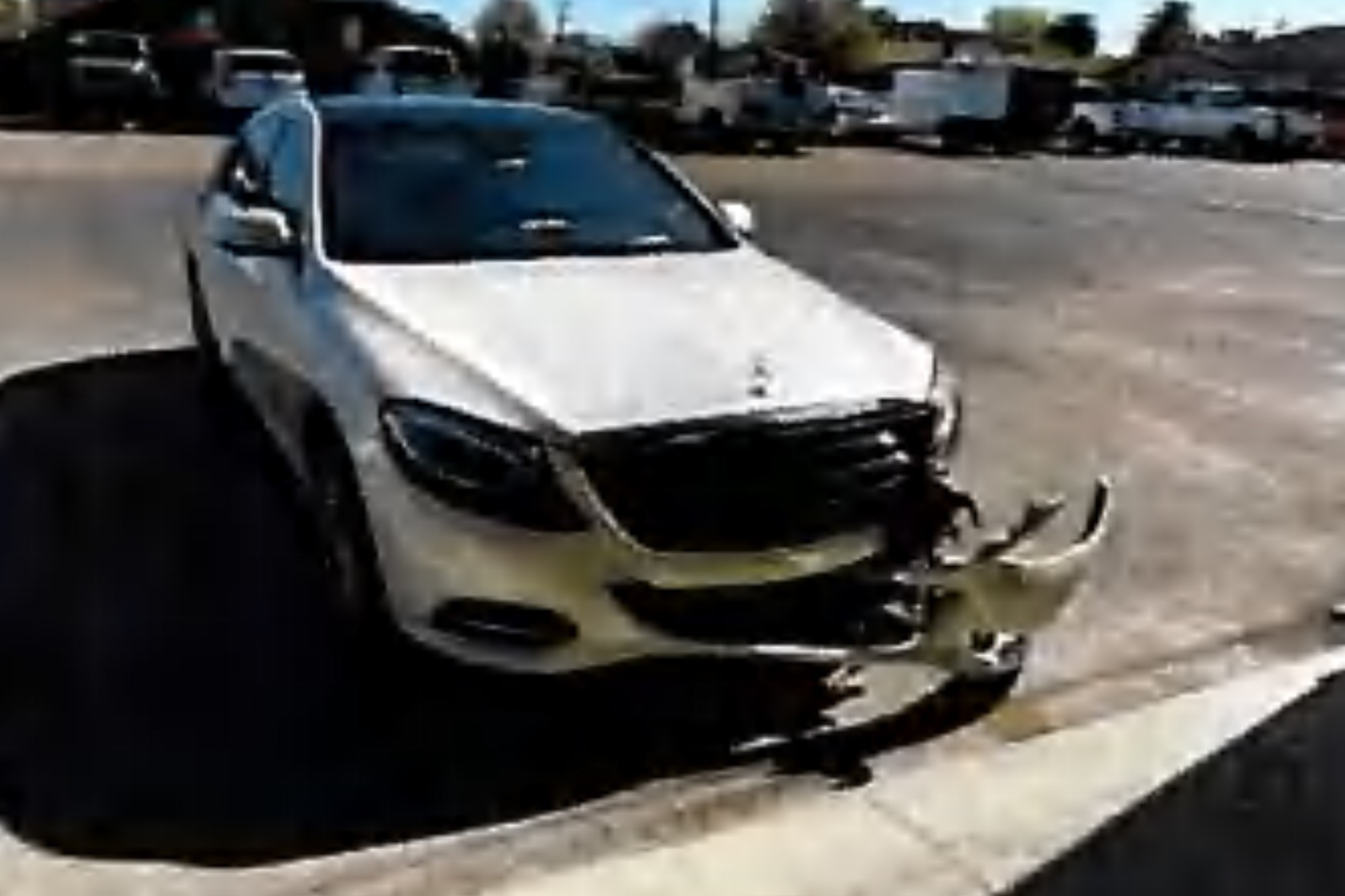 A white Mercedes SUV has front-end damage with its detached bumper hanging off, parked in an outdoor lot with other vehicles in the background.