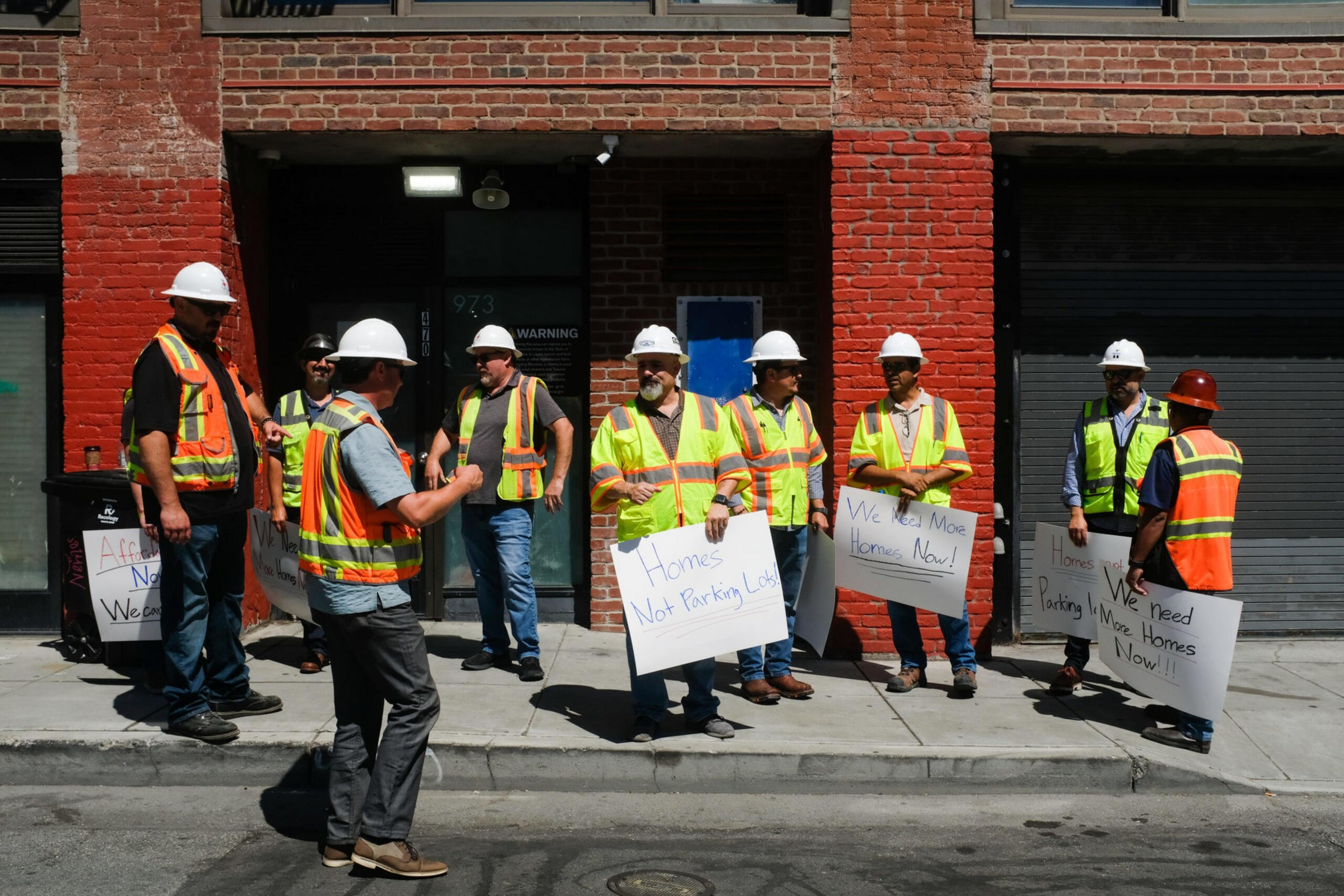 Nine construction workers in safety vests and helmets hold signs demanding more homes instead of parking lots in front of a brick building.