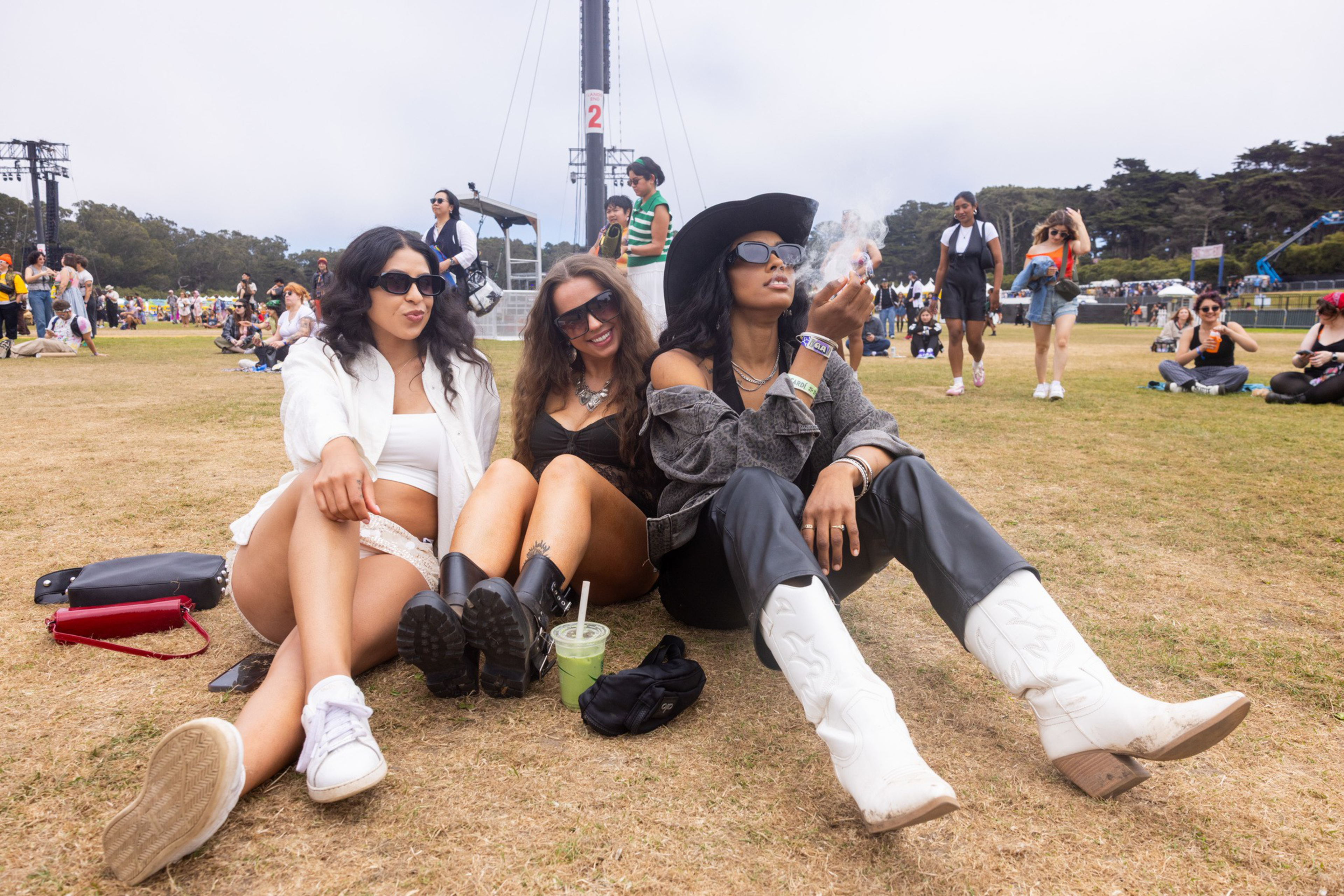 Three women sit on grass at an outdoor event, wearing sunglasses and casual festival attire, with other people and festival structures in the background.