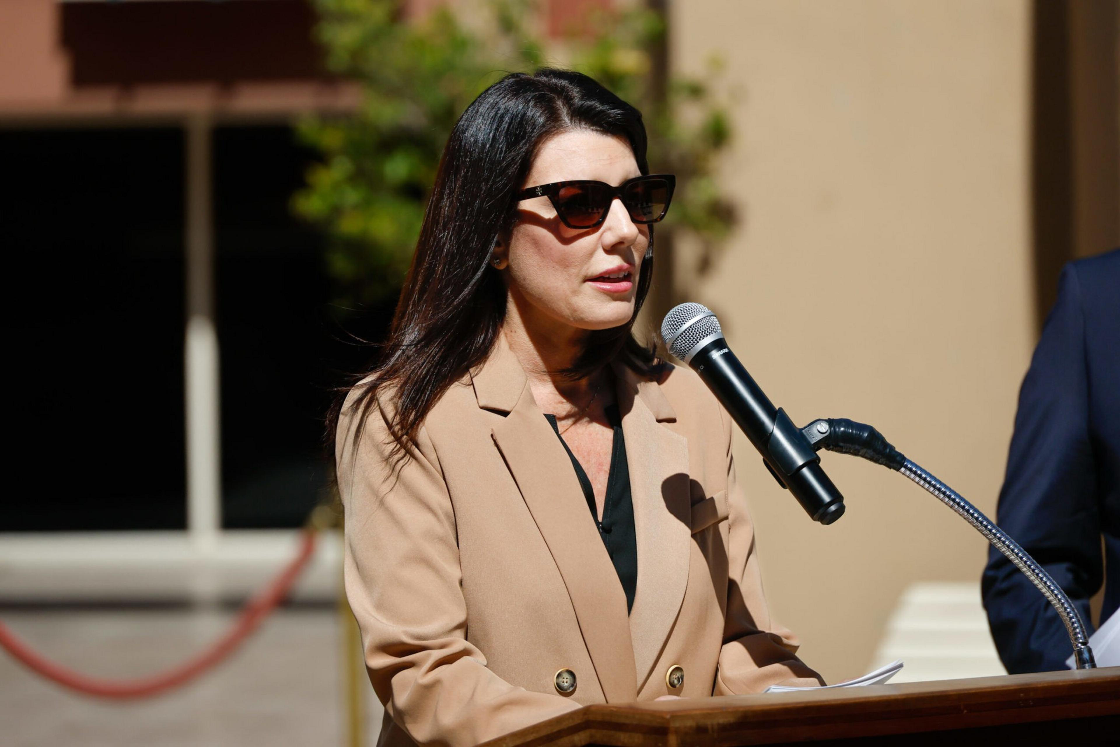 A woman wearing sunglasses and a tan blazer speaks into a microphone at a podium outdoors, with a blurred background of a building and greenery.