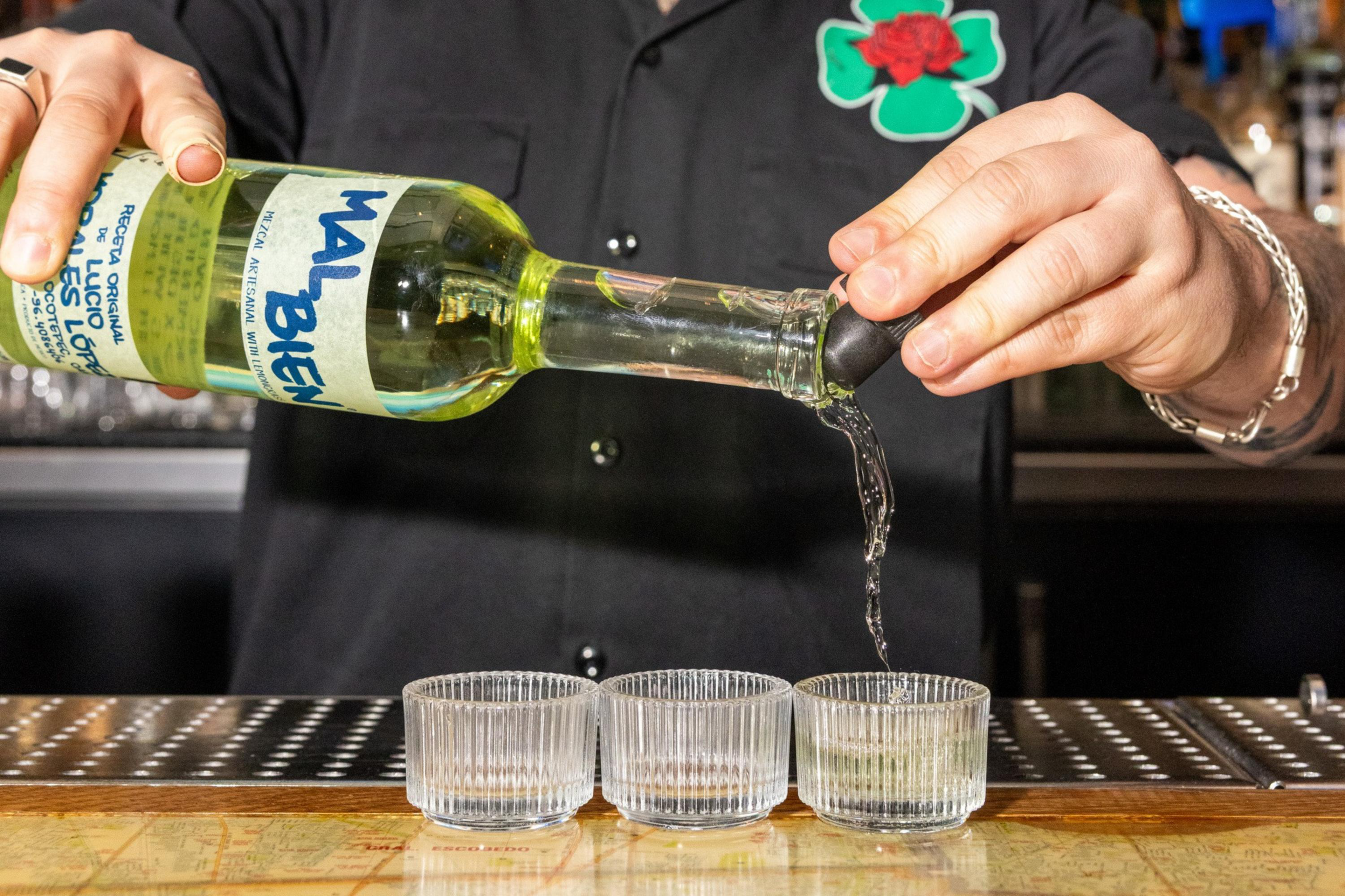A person pours clear liquid from a Mal Bien bottle into three small ribbed glasses on a bar counter.