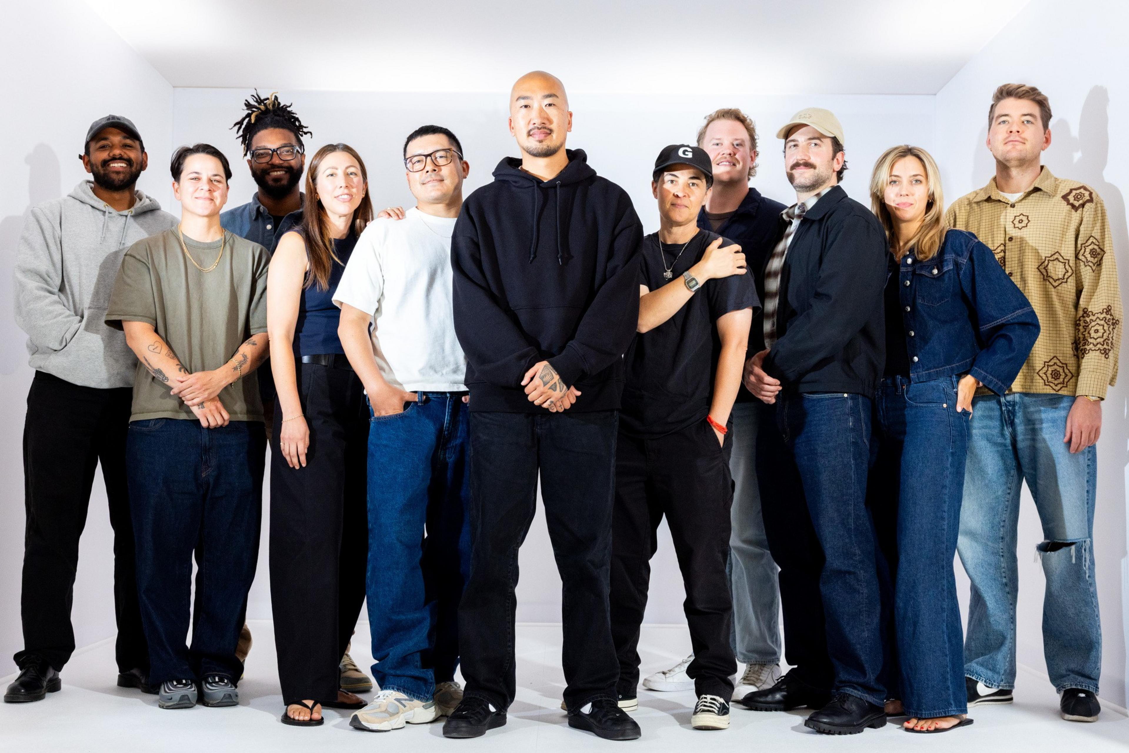Eleven diverse people stand closely together against a white background, dressed casually in jeans, hoodies, jackets, and sneakers, smiling or neutral.