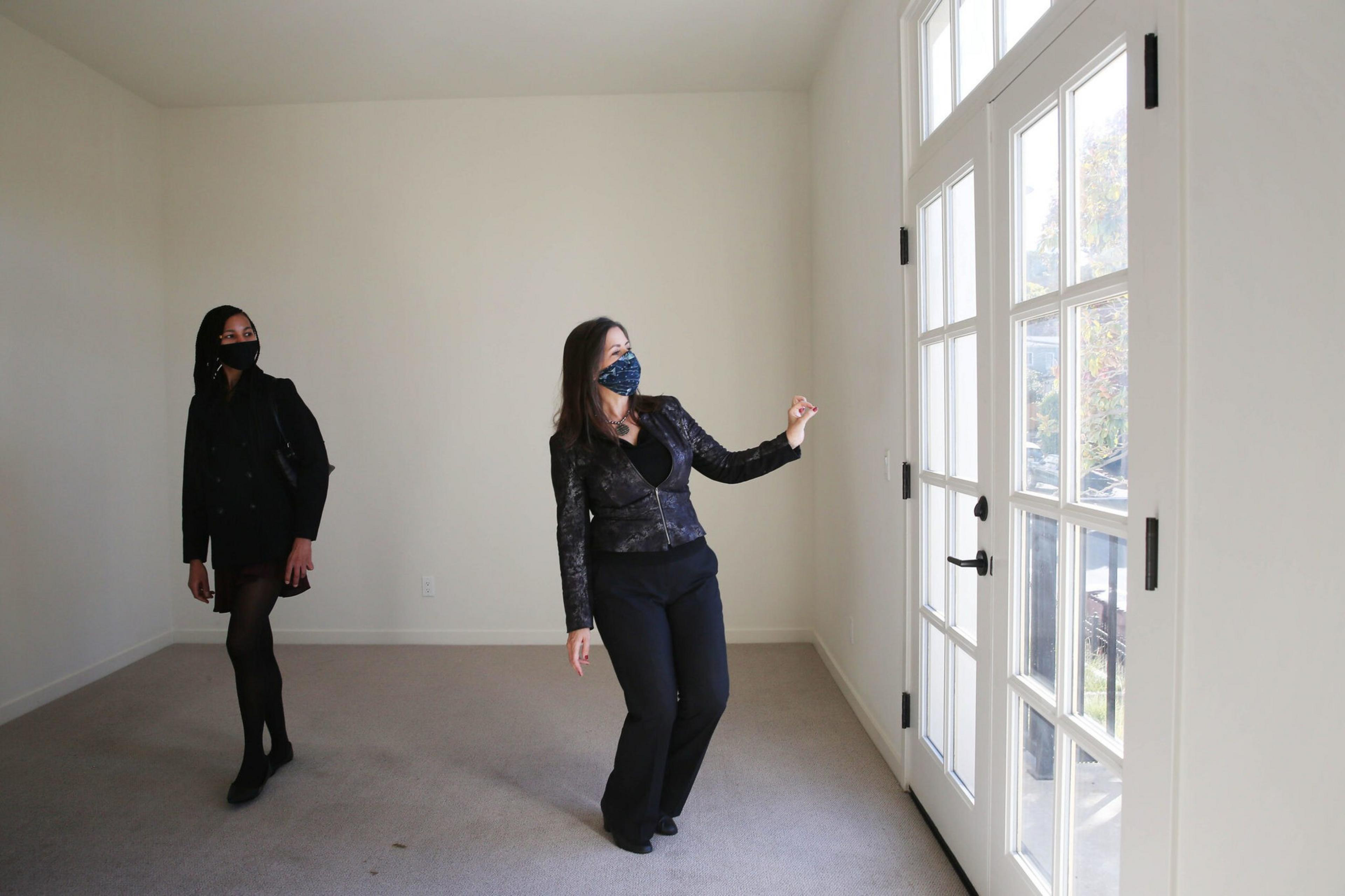 Two women wearing face masks stand in an empty room with light gray carpet, near large white-framed glass doors letting in daylight.