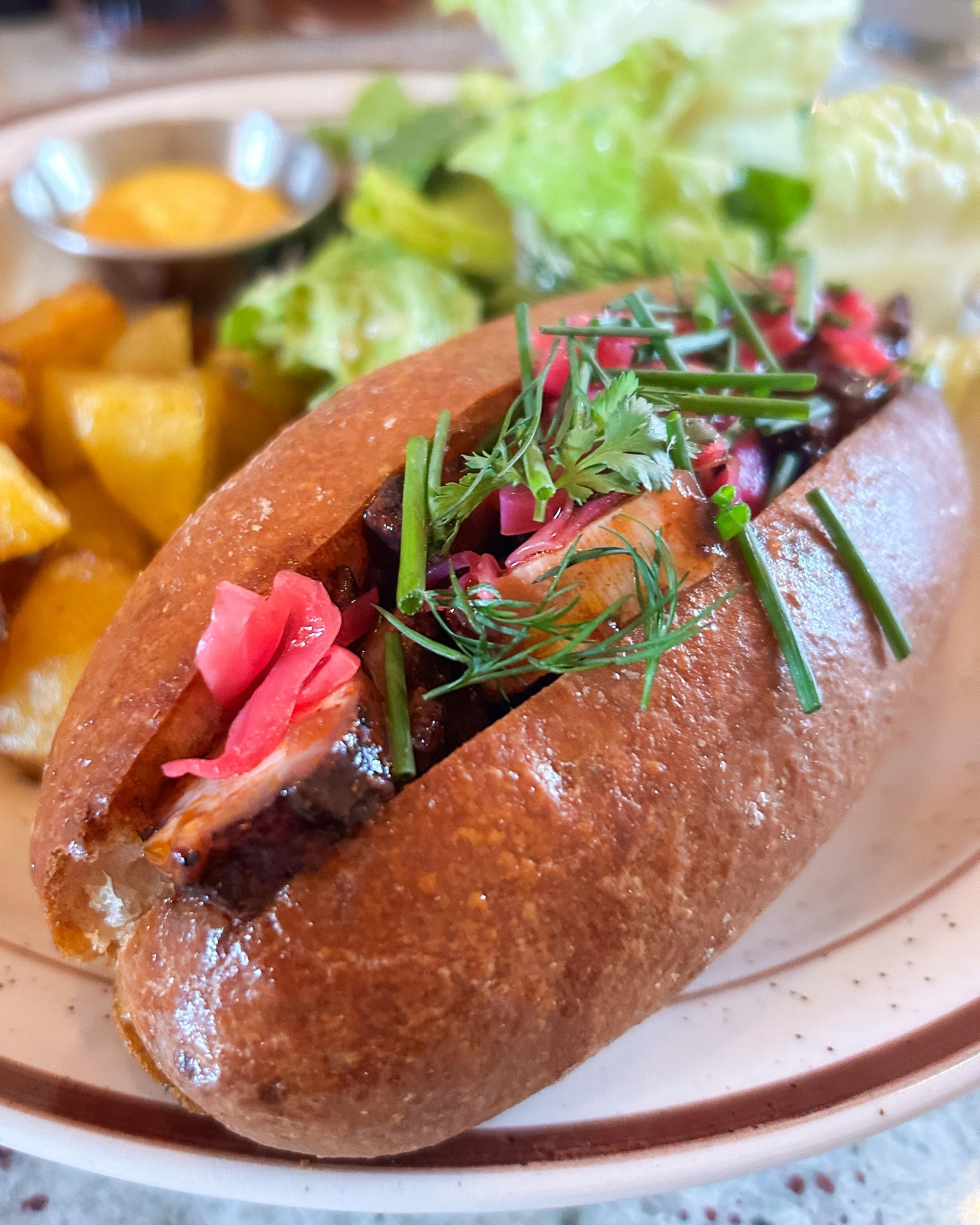 A gourmet hot dog is topped with chopped herbs and pickled onions, served on a plate with roasted potatoes, lettuce, and a small dish of sauce.