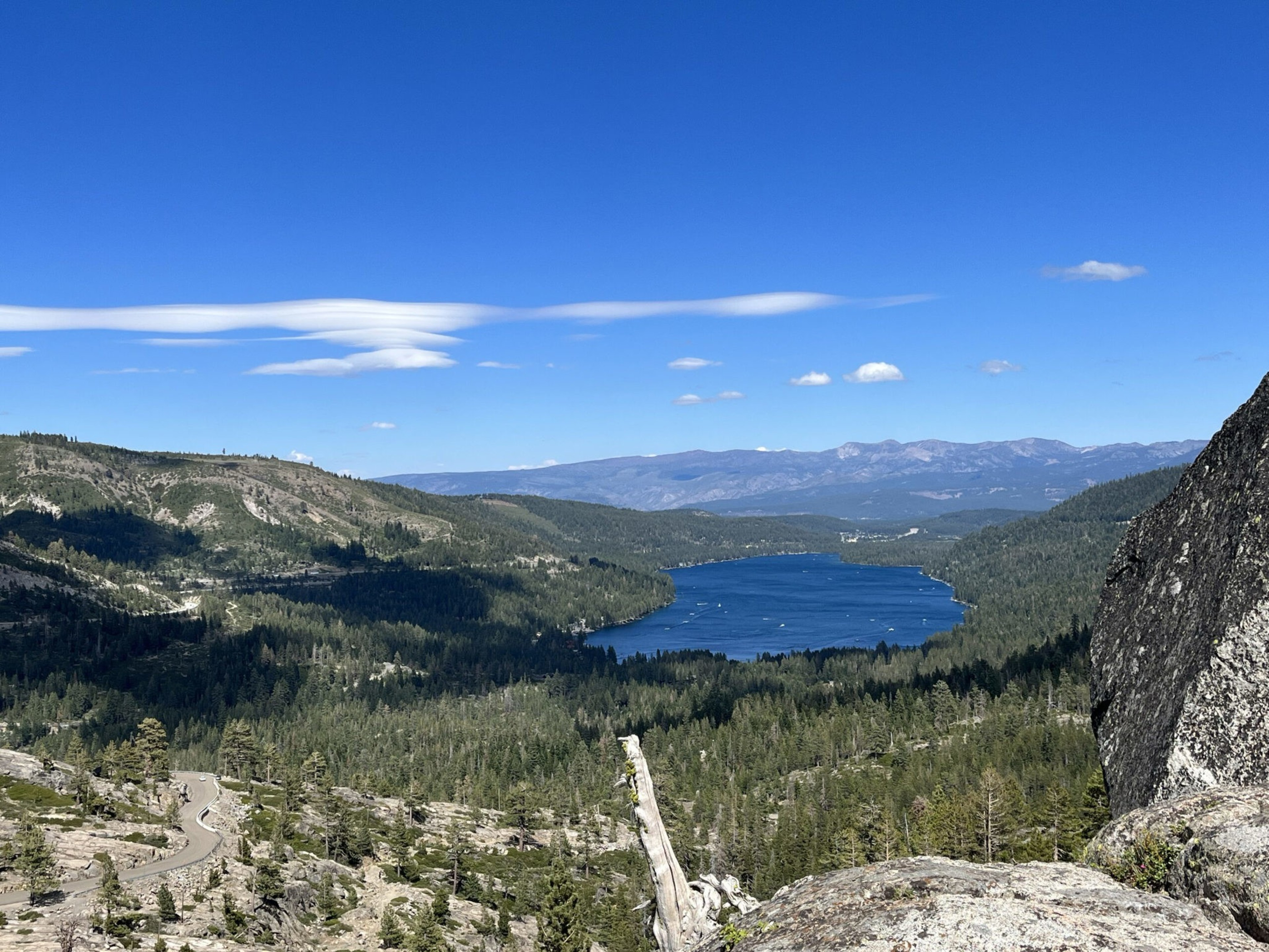 A blue lake surrounded by dense green forest and mountains under a clear blue sky with a few scattered clouds is visible.