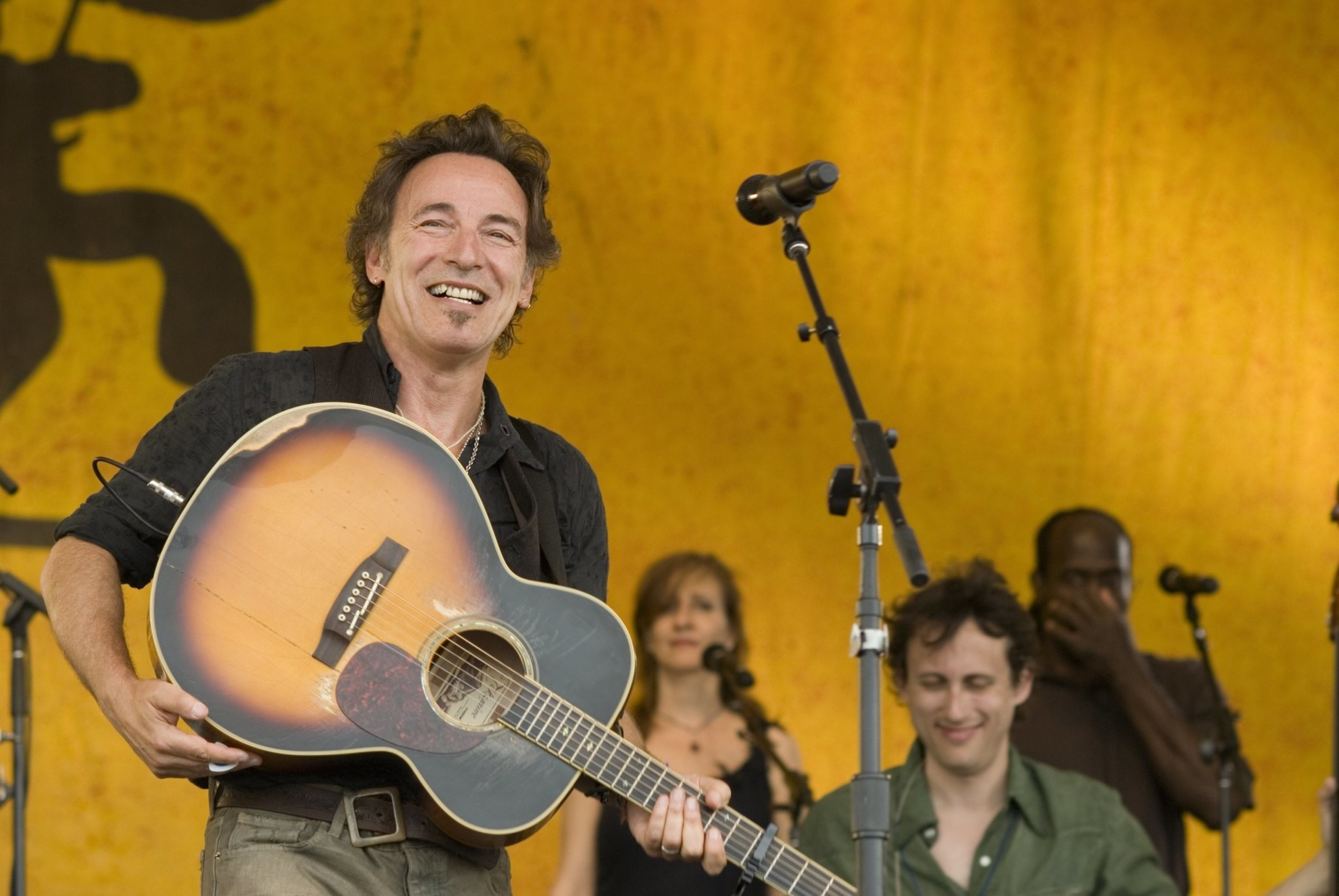 A person is joyfully holding an acoustic guitar on stage with three people in the background. The backdrop is yellow, and microphones are present.