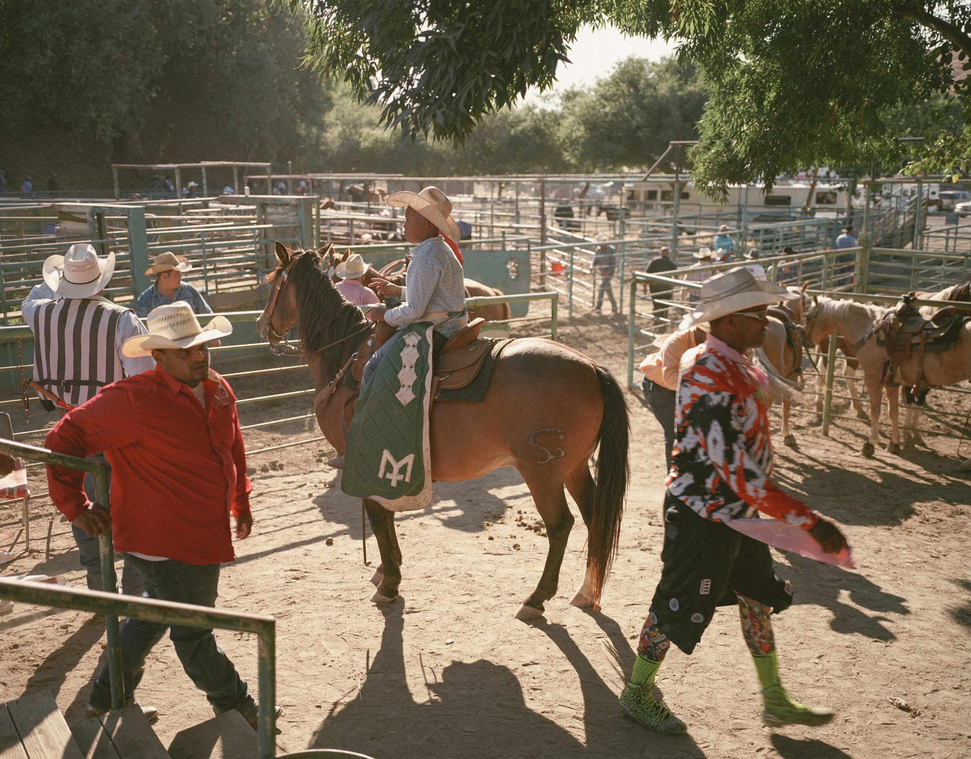A group of people in cowboy hats are in a sunny rodeo arena. One person rides a horse, while others walk or stand, all dressed in vibrant cowboy attire.