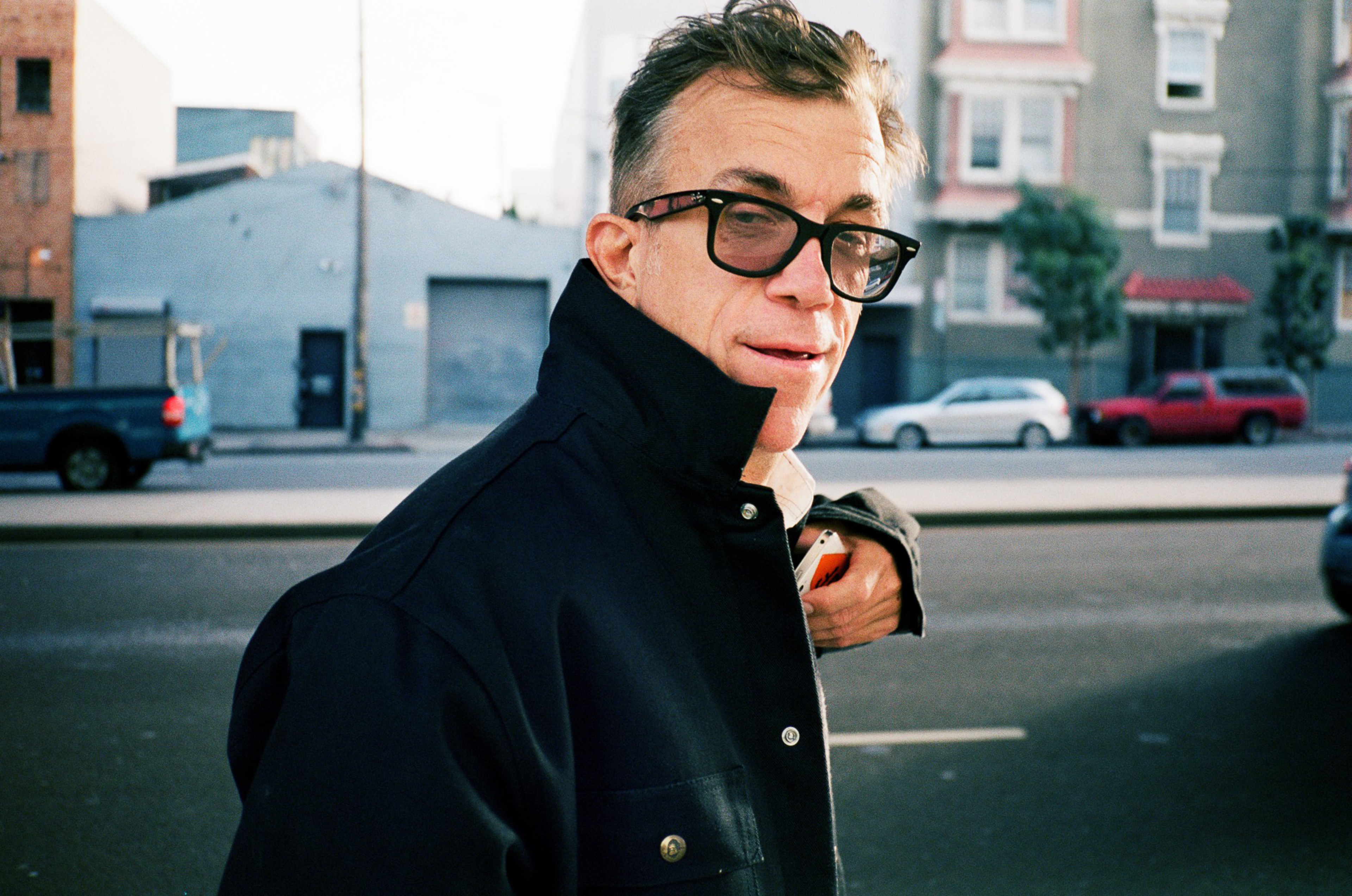 A middle-aged man with short hair and black glasses wears a dark jacket and looks back while standing on a city street with cars and buildings behind him.