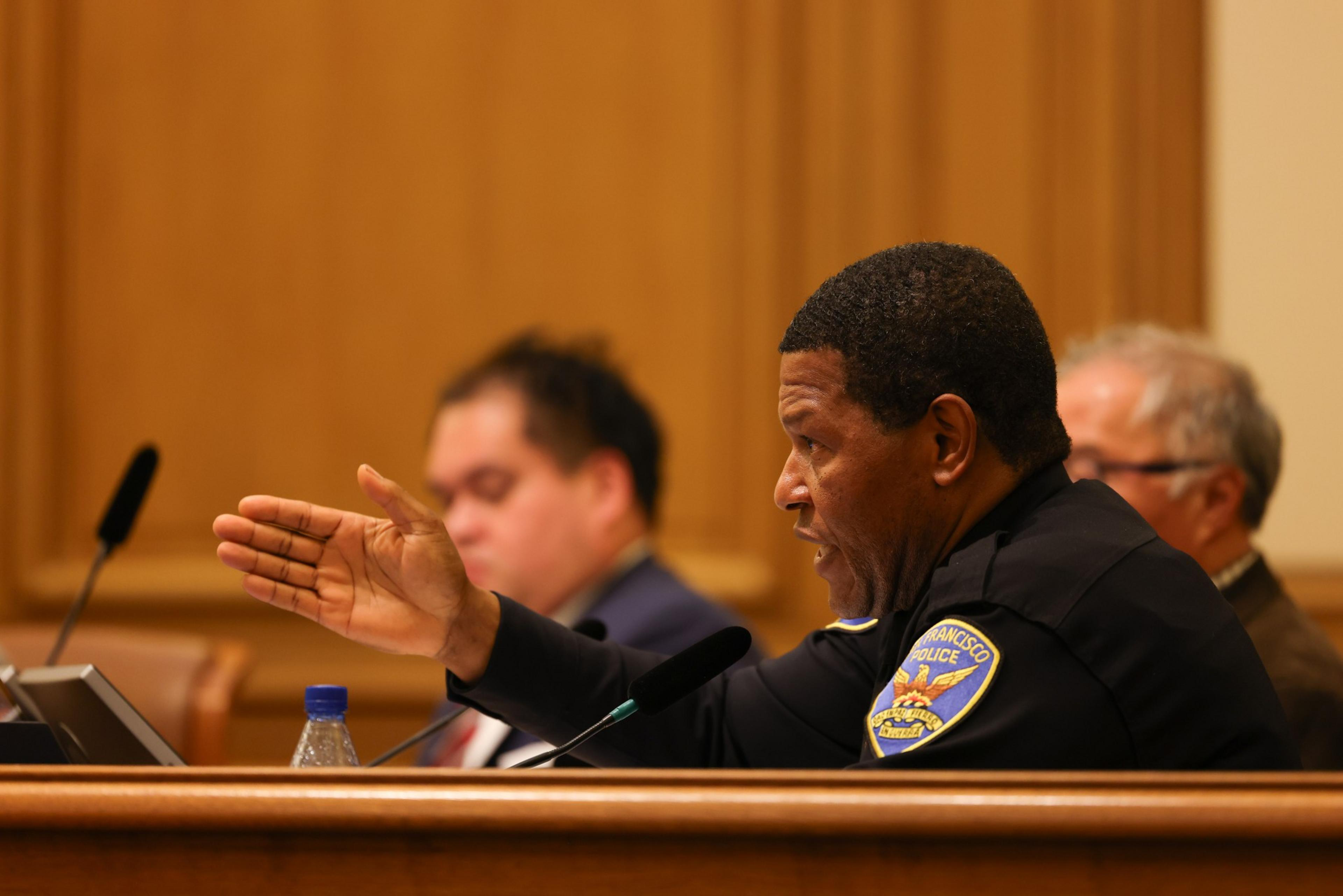 A man in a police uniform speaks at a meeting, gesturing with his hand. Two other people sit nearby, one blurred in the background, with a water bottle visible.