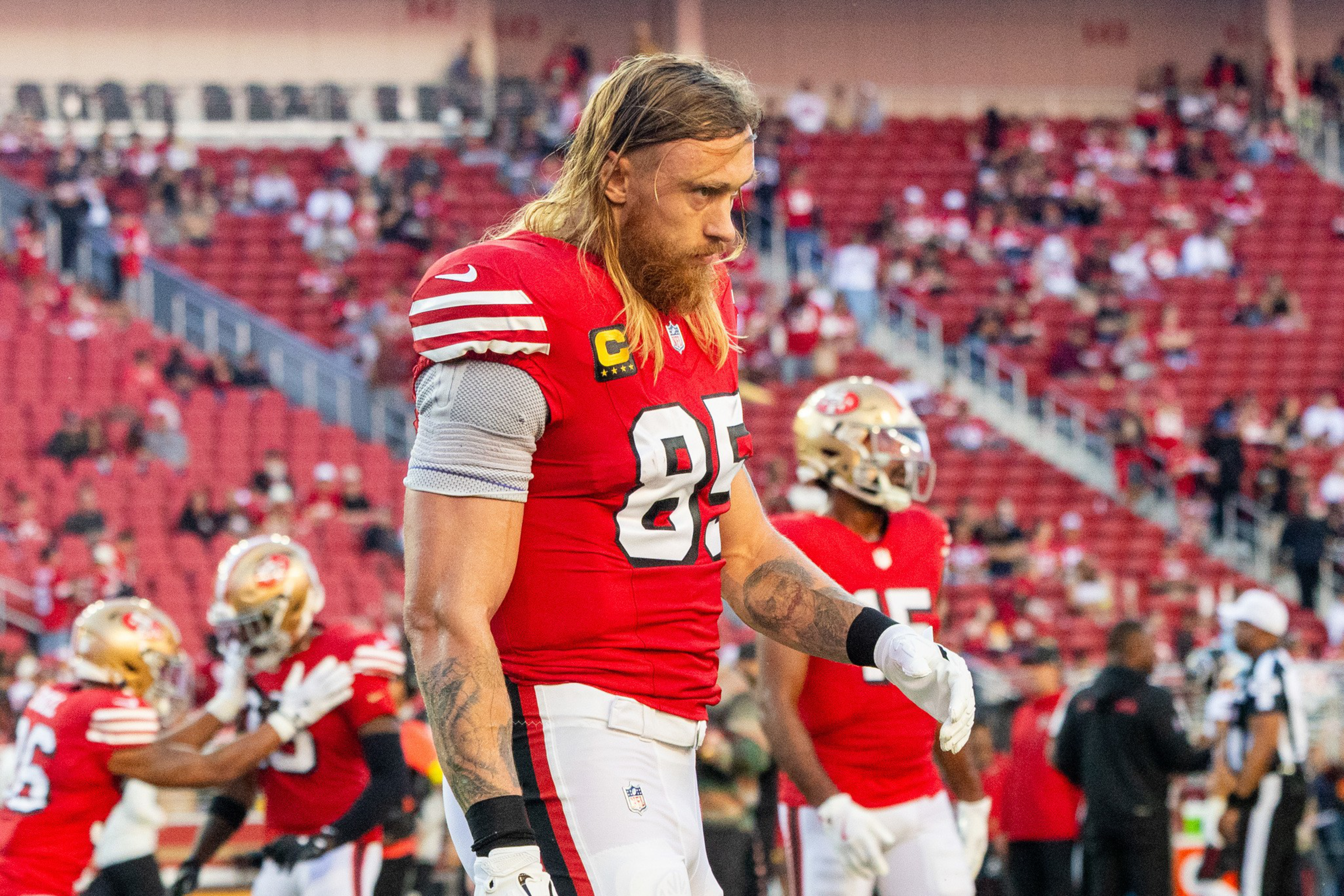 A football player with long hair and a beard in a red jersey number 85 looks down with focus while warming up on the field before a game.