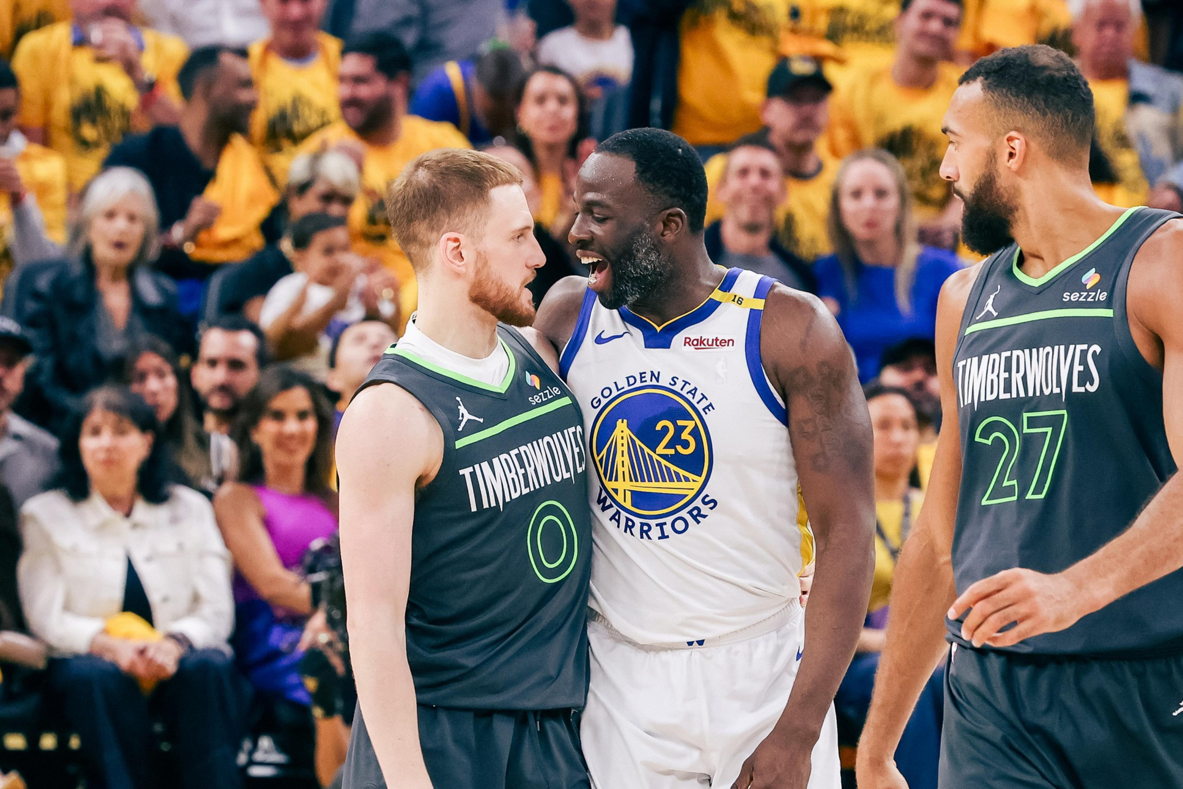 A basketball player from the Warriors shouts in the face of a Timberwolves player during a game, while another Timberwolves player looks on. Fans fill the background.