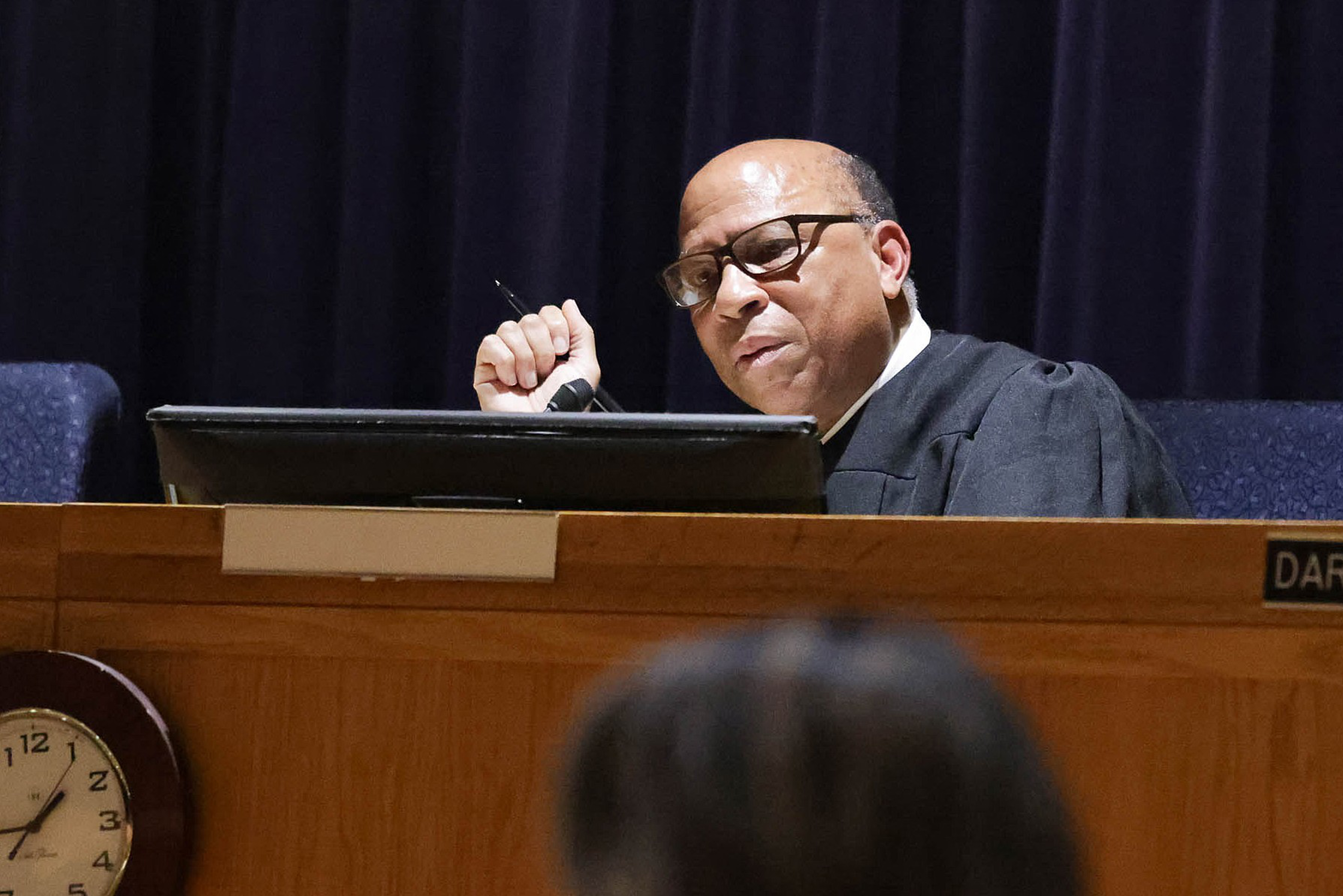 A man in judicial robes sits behind a bench with a microphone, pen in hand, and a clock in the foreground.