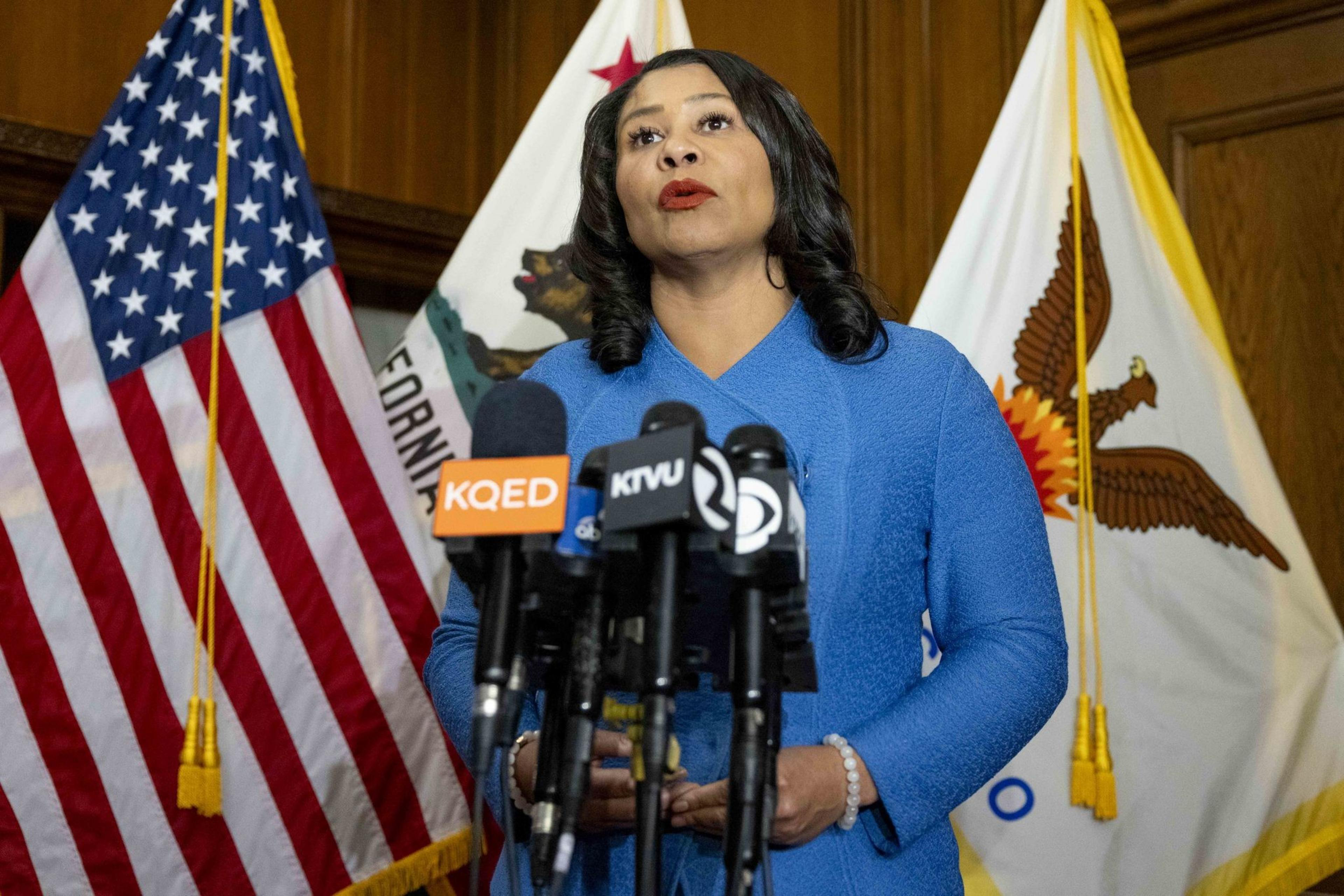 A woman in a blue outfit speaks at a podium with multiple microphones. Behind her are U.S., California, and city flags.