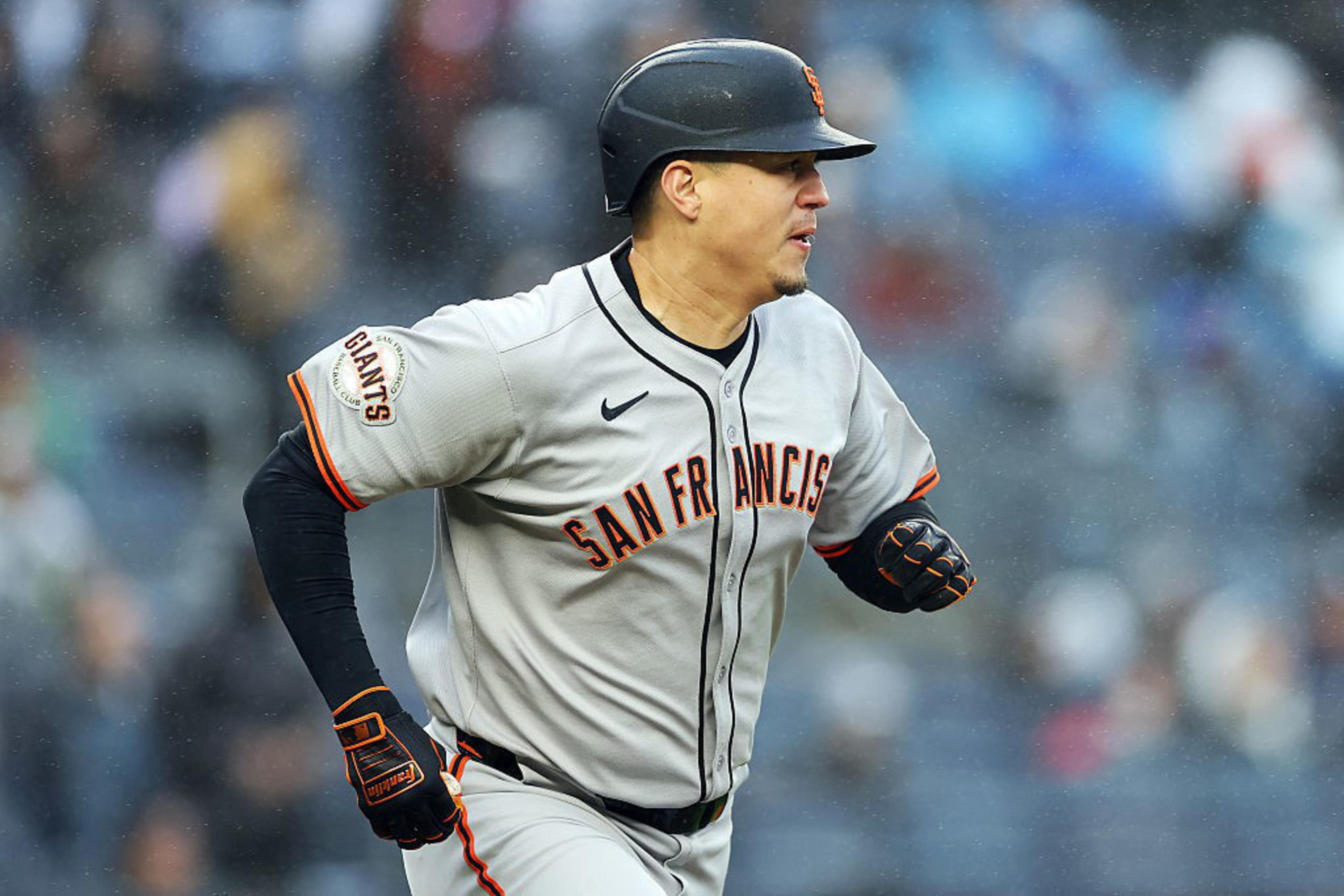 A baseball player in a gray "San Francisco" jersey runs, wearing a black helmet and gloves. The background shows a blurred crowd, suggesting a game in progress.