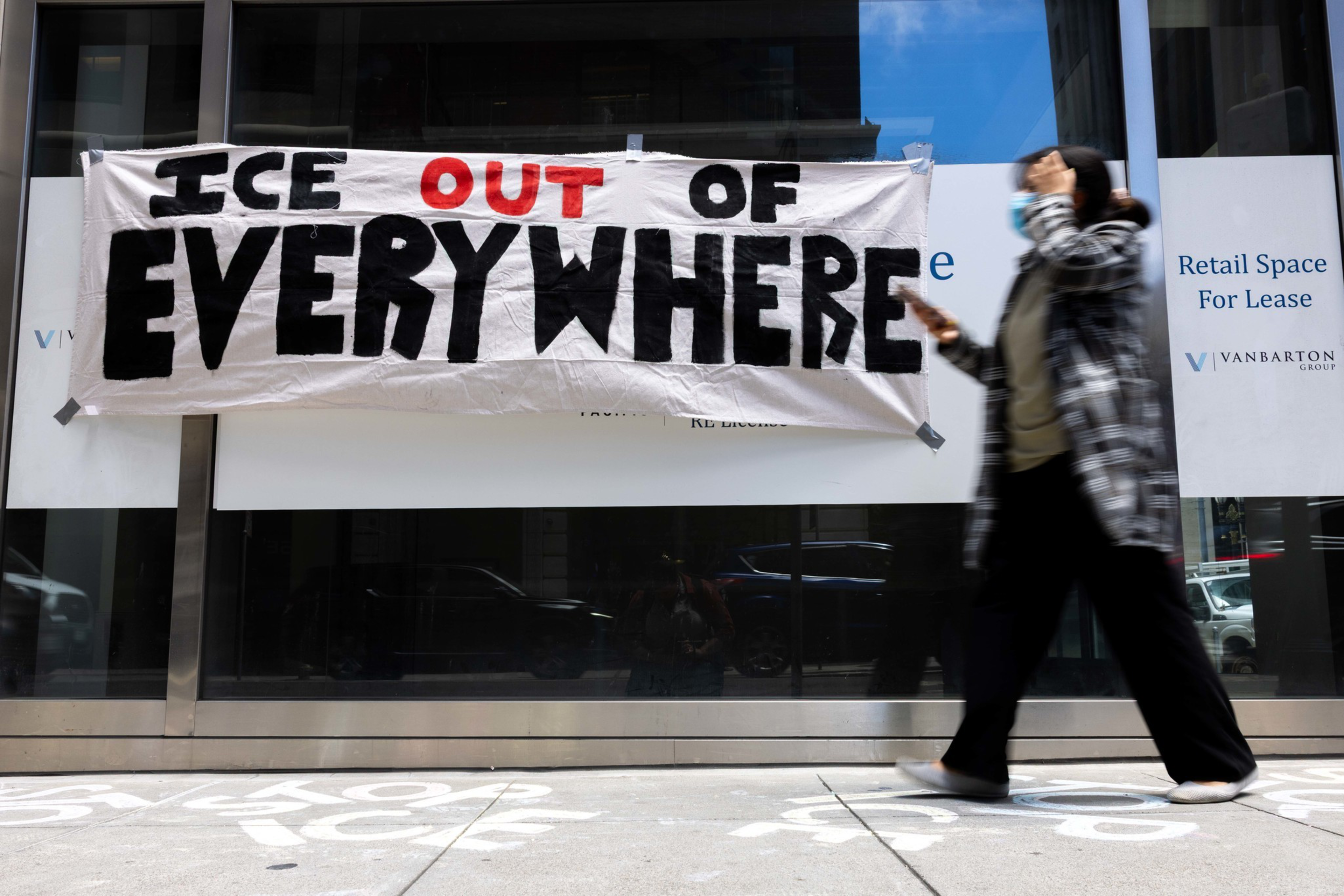 A banner reads &quot;ICE OUT OF EVERYWHERE&quot; in black and red letters on a building. A person in a mask and checkered coat walks by, holding a phone.