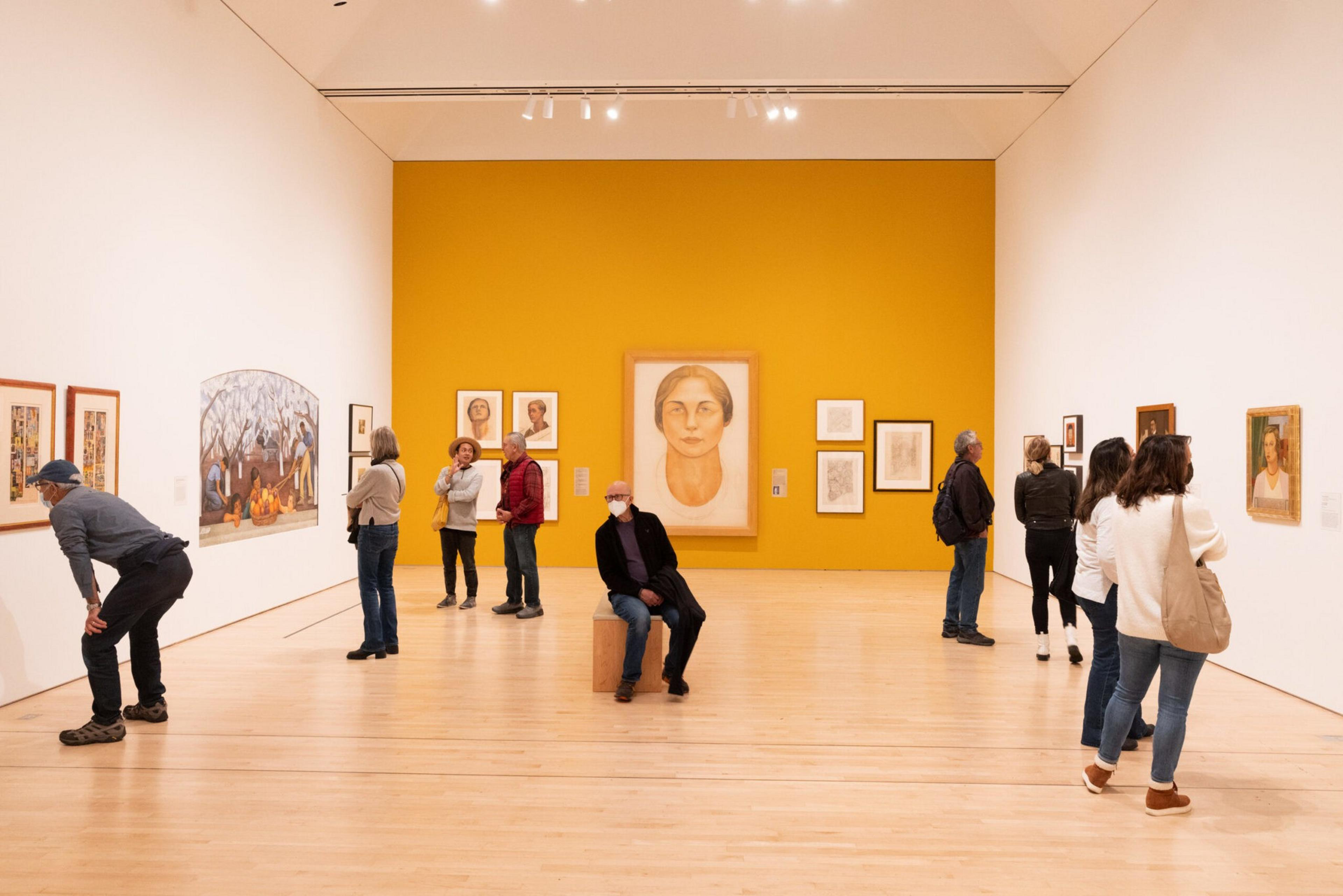 People observe various framed artworks on white and mustard-yellow walls in a spacious, well-lit modern art gallery with light wood flooring.