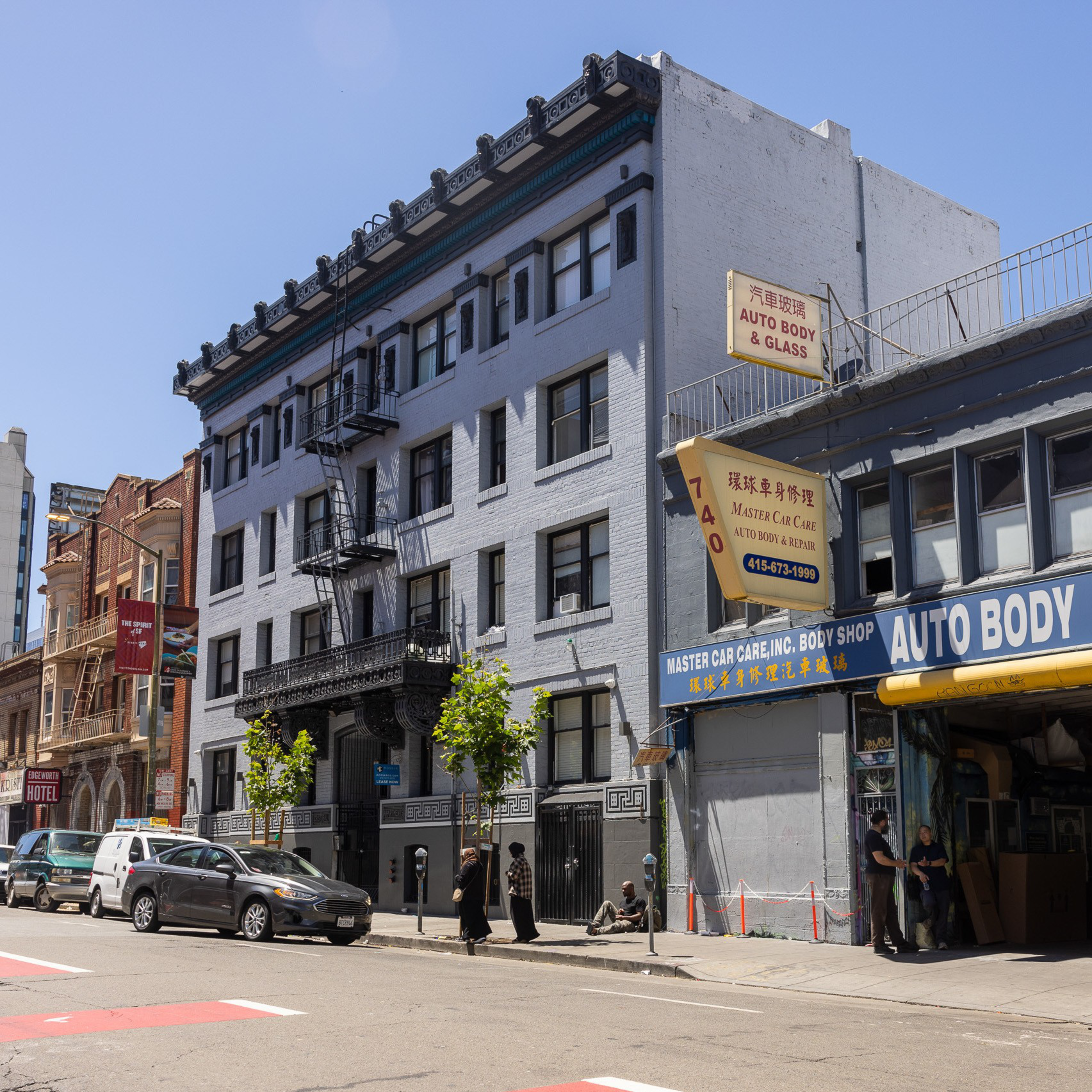 The image shows a street with a tall grey building, a red brick building beside it, and an auto body shop. People and parked cars are on the street below.