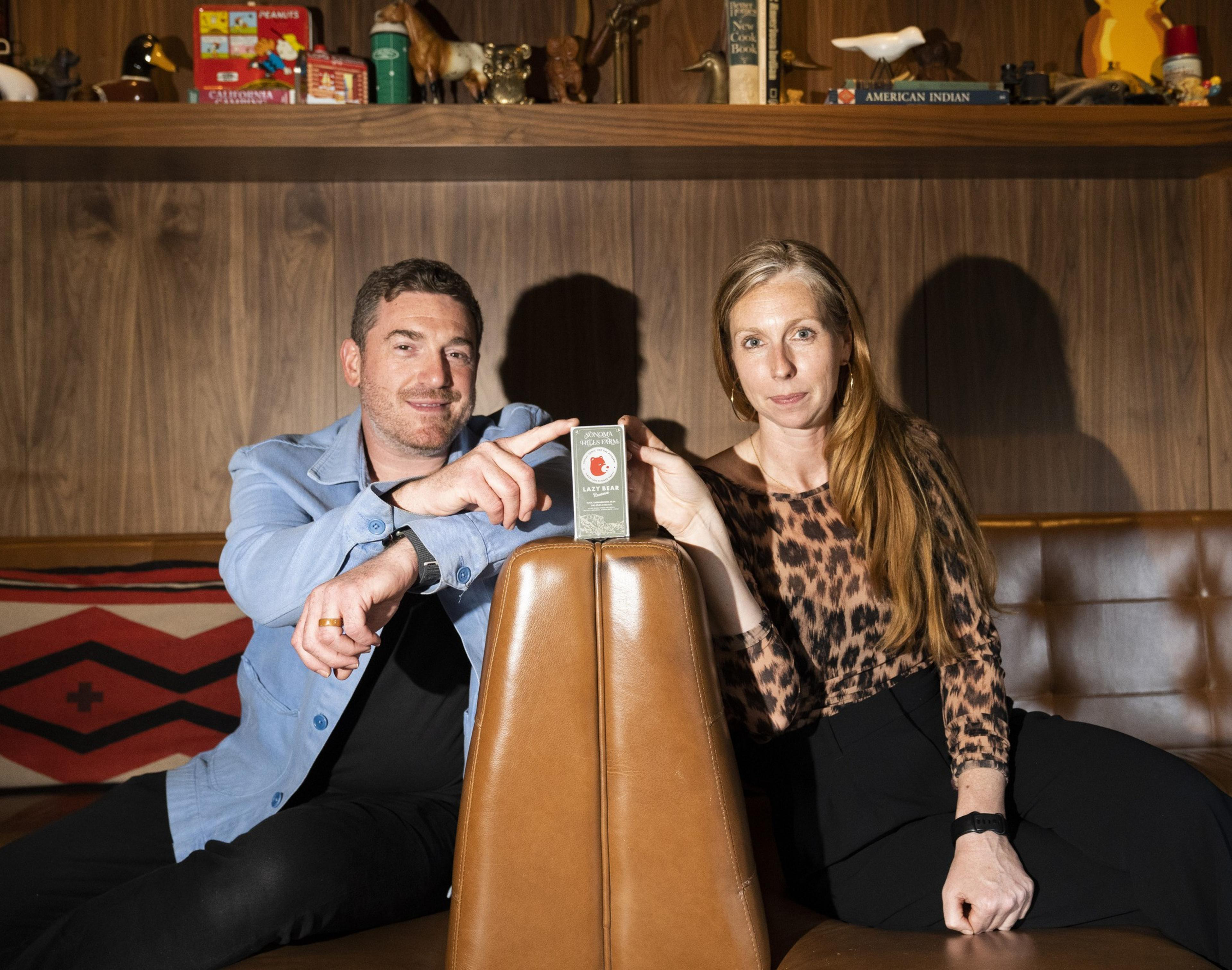 A man in a light blue shirt and a woman in a leopard-print top are seated on a leather sofa, pointing at a small box. Books and trinkets are on a shelf behind them.