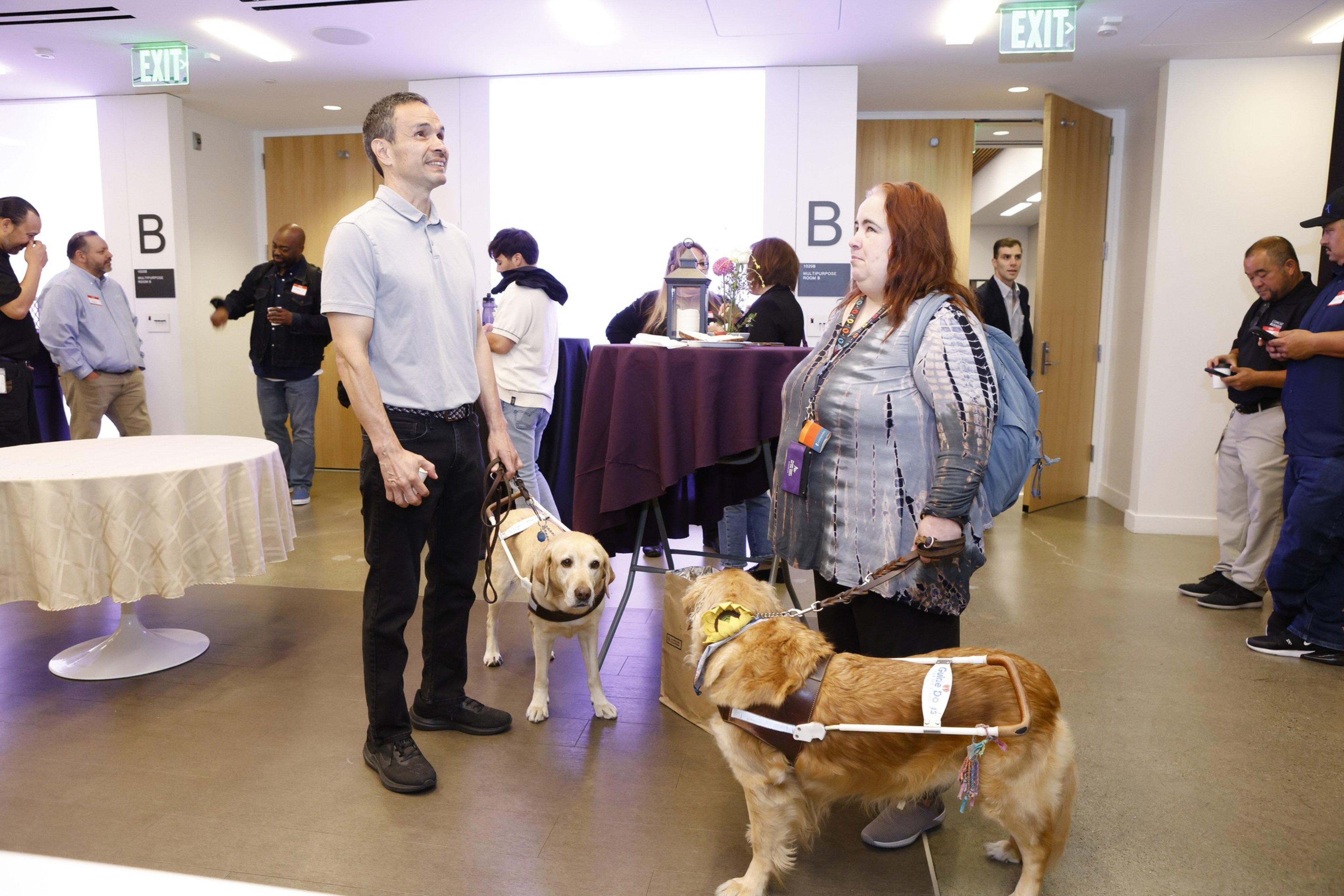 Two people with guide dogs converse in a well-lit room with eight other people chatting. The guide dogs are a Labrador and a Golden Retriever.