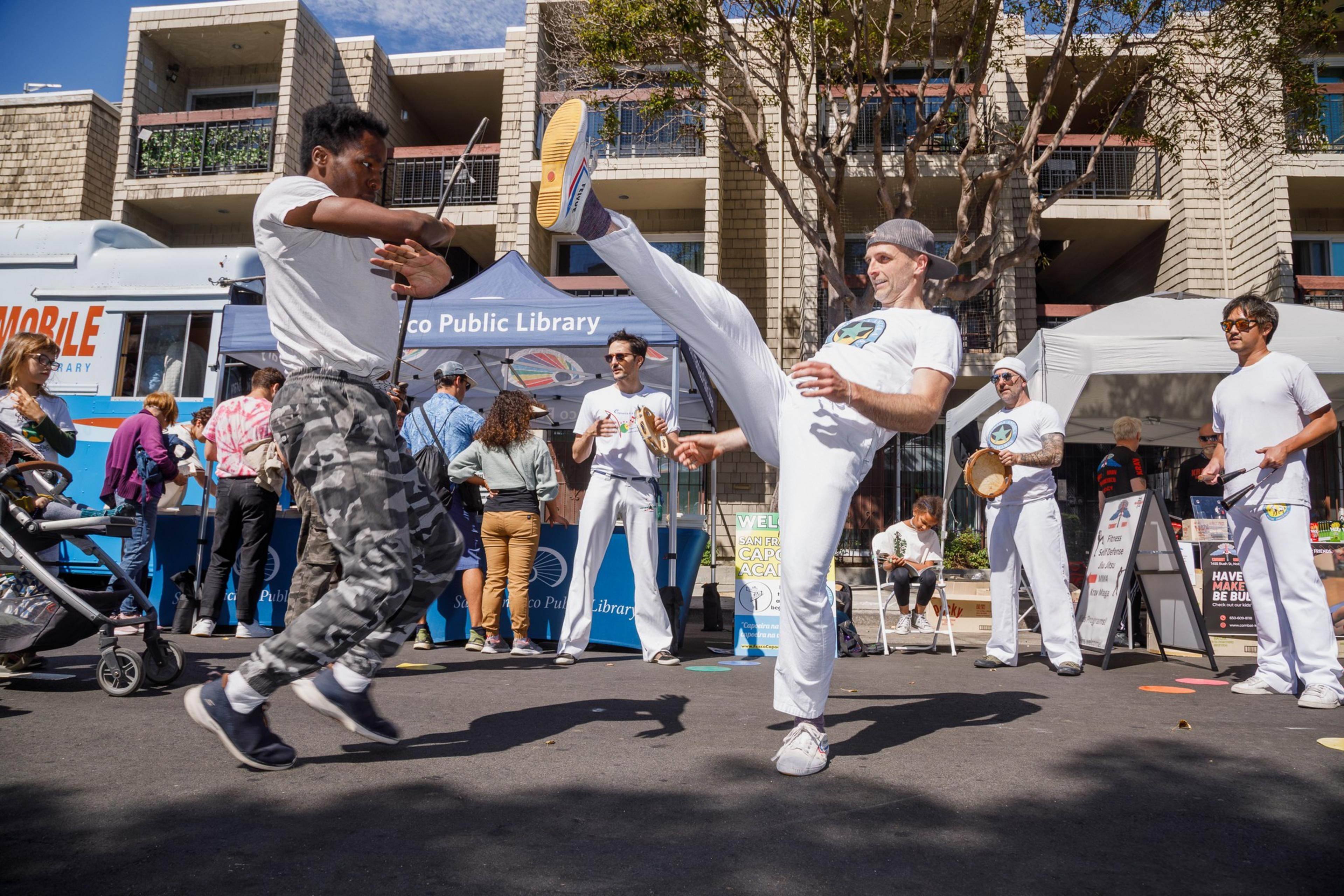 People perform Capoeira, a Brazilian martial art, on a sunny street with onlookers and a library mobile in the background.