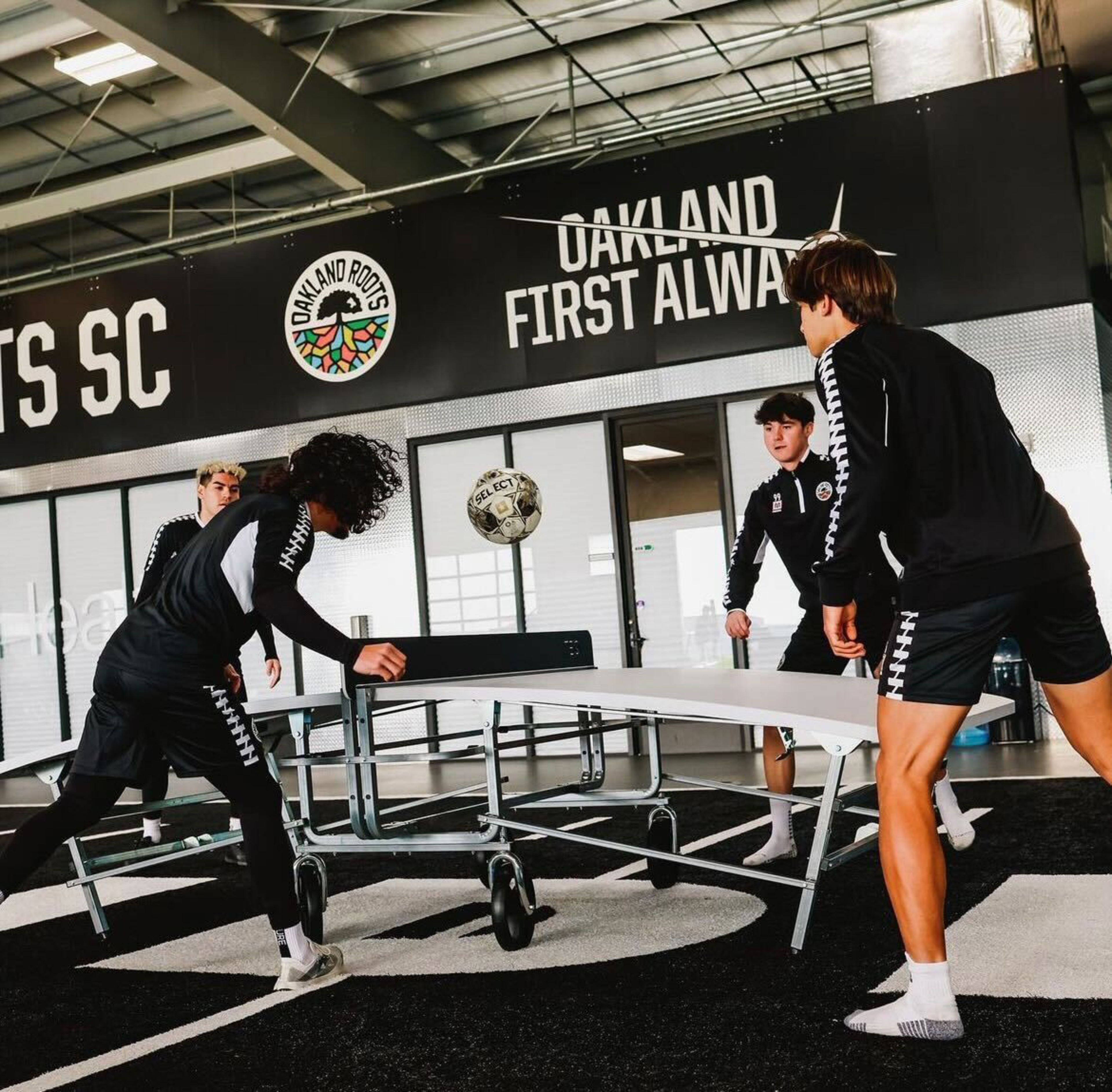 Four young men in matching black sportswear play a game with a soccer ball over a ping pong table indoors under a sign reading "Oakland First Always."