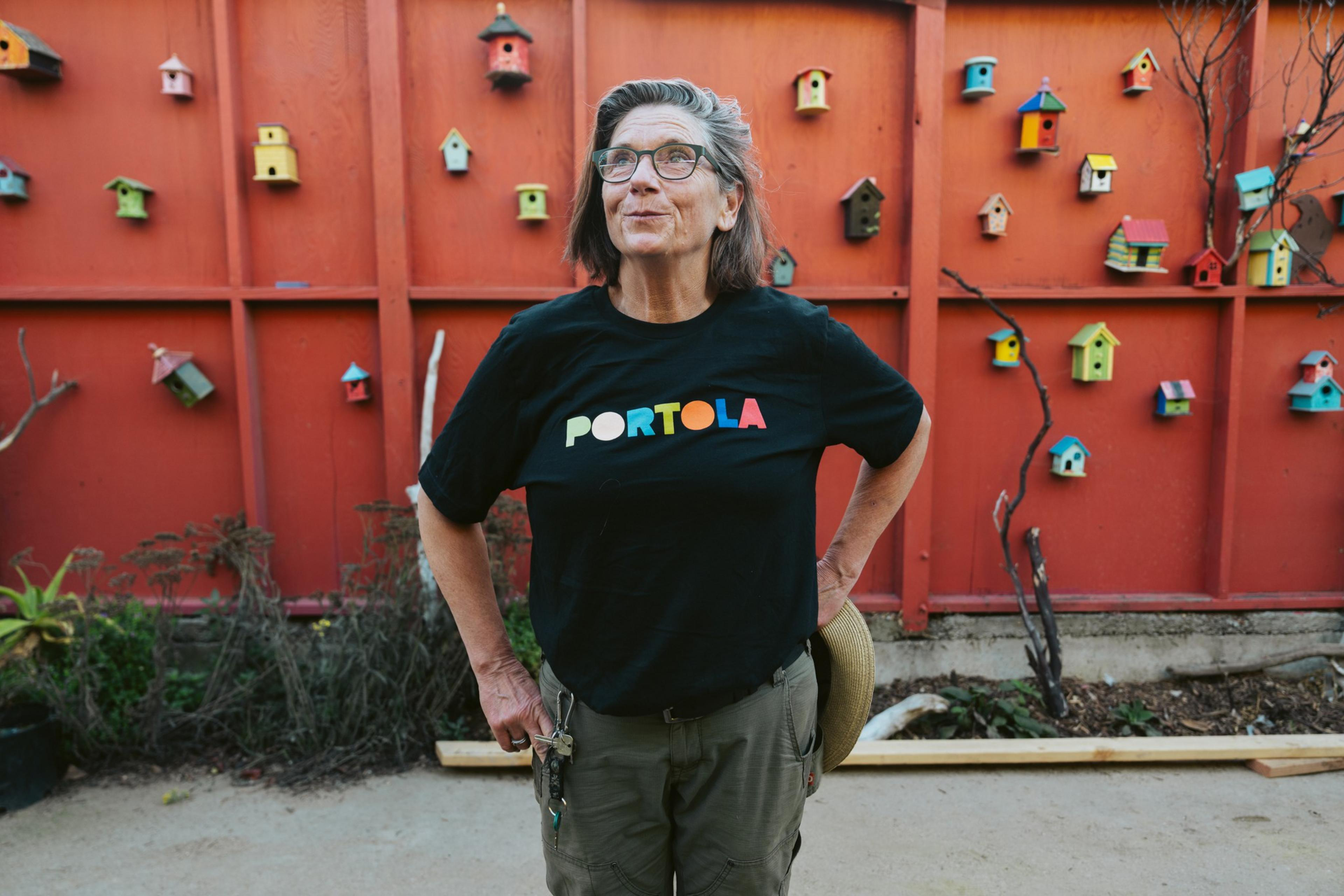 A person with gray hair and glasses stands in front of a red wall adorned with colorful birdhouses, wearing a black &quot;Portola&quot; T-shirt and green pants.